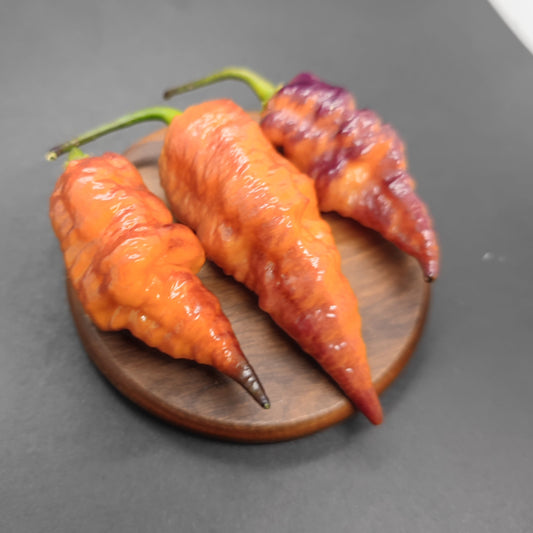 Three wrinkled chili peppers—two orange-red and one partially purple—rest on a small wooden plate against a dark background, highlighting PepperMerchant.net’s PM030 - Jays x Pink x Lemon Ghost variety.