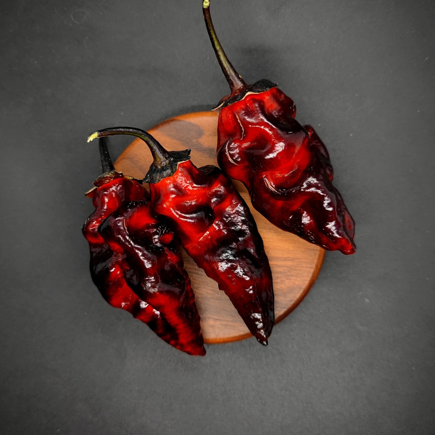 Three BBG7 Alchemy - Dark Red Pheno peppers from PepperMerchant.net, with wrinkled skin and green stems, rest on wood against a black backdrop—highlighting their fiery look and ideal for those seeking unique superhot pepper seeds.
