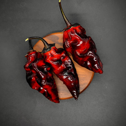 Three BBG7 Alchemy - Dark Red Pheno peppers from PepperMerchant.net, with wrinkled skin and green stems, rest on wood against a black backdrop—highlighting their fiery look and ideal for those seeking unique superhot pepper seeds.