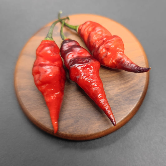 Three wrinkled red chili peppers with stems rest on a round wooden surface, set against a dark background—ideal inspiration for growers looking for unique seeds like PepperMerchant.net’s Black Chupetinho Long Cross.