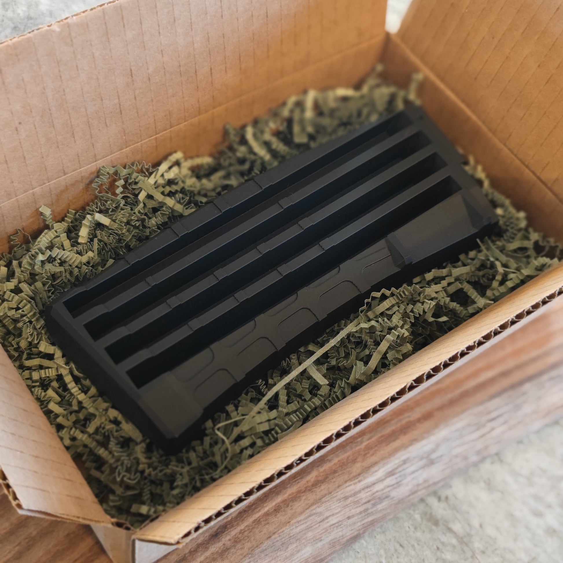 The GEN4 CoinMag - Black from coin-mag.net, a black rectangular grooved challenge coin holder, is nestled in shredded packing paper inside an open brown cardboard box on a wooden surface.