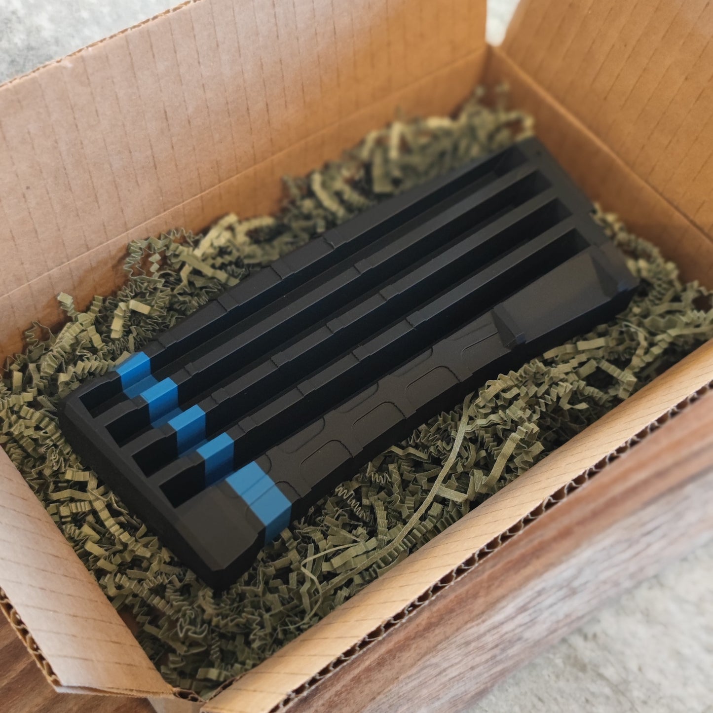 The GEN4 CoinMag - Blue Line by coin-mag.net, a black rectangular challenge coin display with blue accents, sits on green shredded packing material inside an open cardboard box.