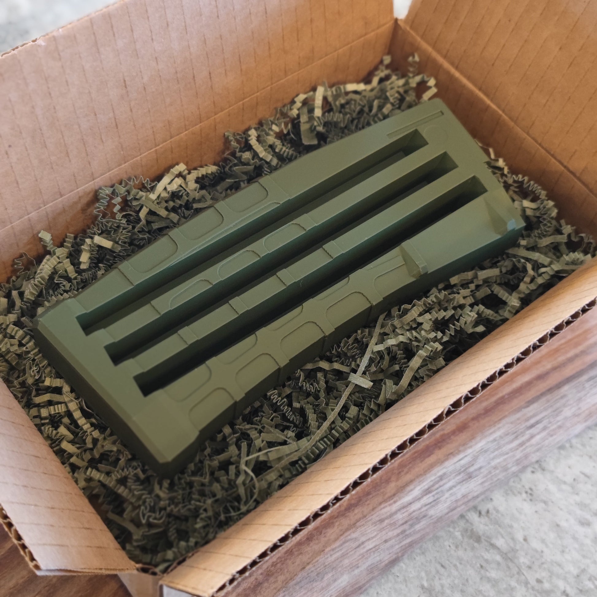 The GEN3 CoinMag - Olive by coin-mag.net, a green rectangular plastic coin holder with grooves and slots, rests in shredded packing material inside an open cardboard box.