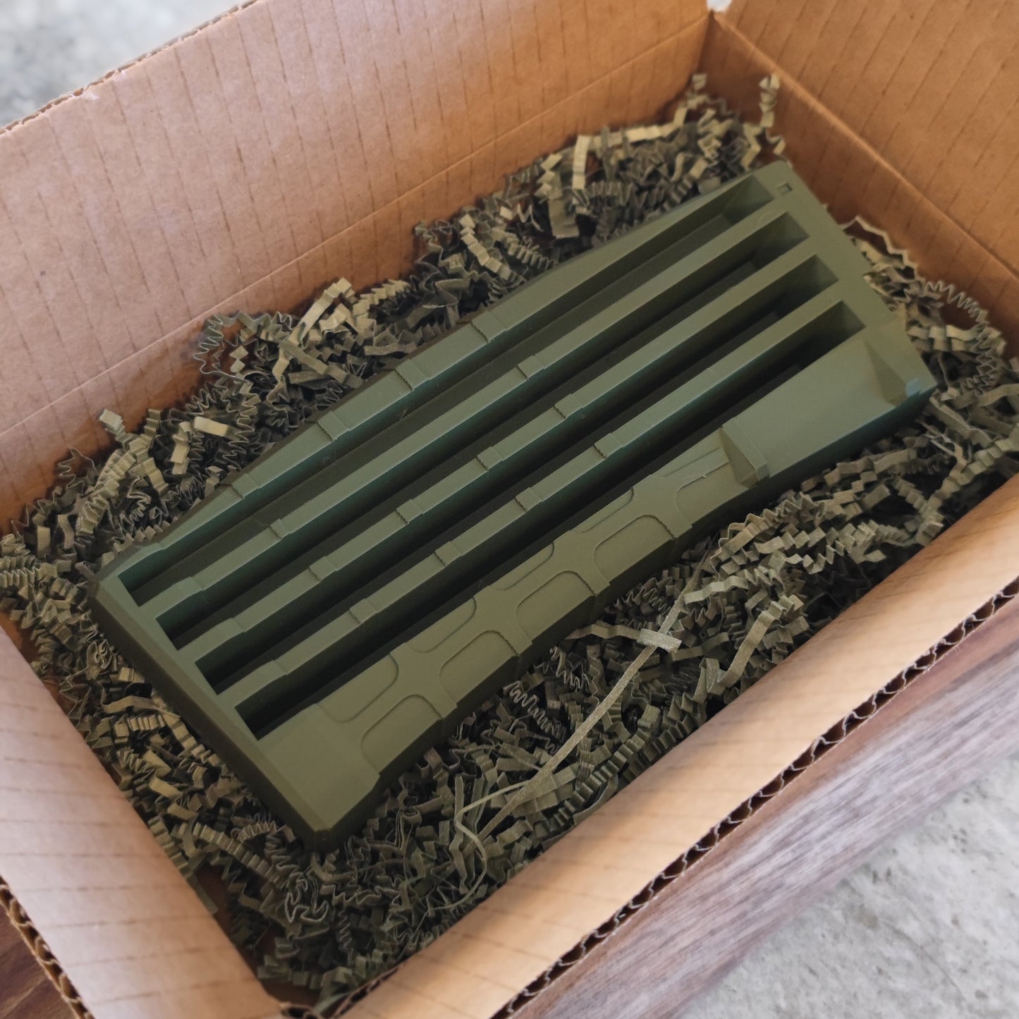 The GEN4 CoinMag - Olive from coin-mag.net, a green plastic vent cover, rests in a cardboard box with crinkled paper next to a sleek magazine-style coin display ready to showcase your favorite coins.