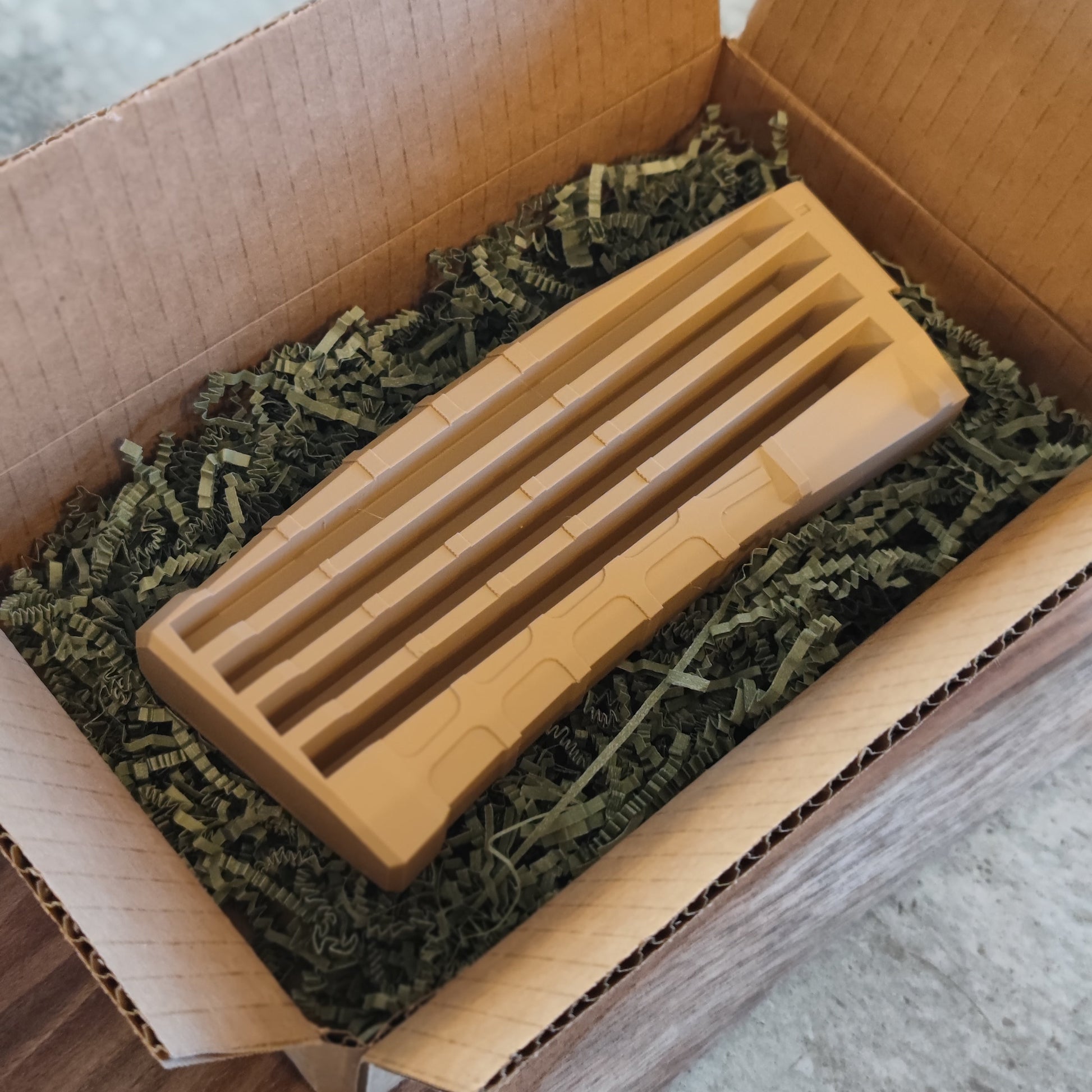 A GEN4 CoinMag - Sand by coin-mag.net, a tan ribbed plastic challenge coin holder, sits on green shredded packing paper inside an open cardboard box on a wooden surface.