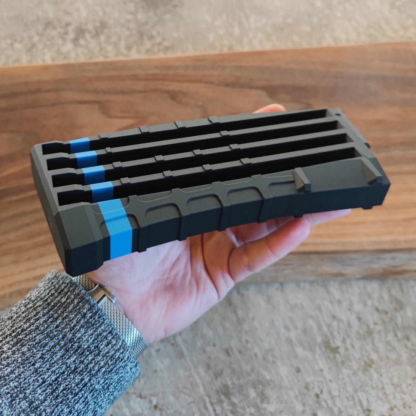A hand holds a GEN4 CoinMag - Blue Line by coin-mag.net, a rectangular black challenge coin holder with grooves and blue accents, against a blurred wood and concrete background.