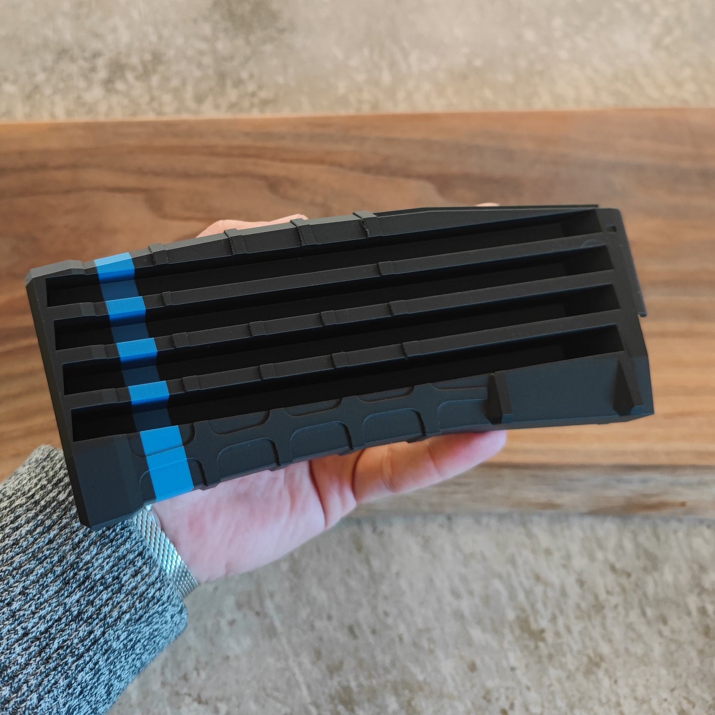 A hand holds the GEN4 CoinMag - Blue Line by coin-mag.net, a black rectangular challenge coin holder with four horizontal slots and two vertical blue stripes, set on a wooden surface with a textured floor background.