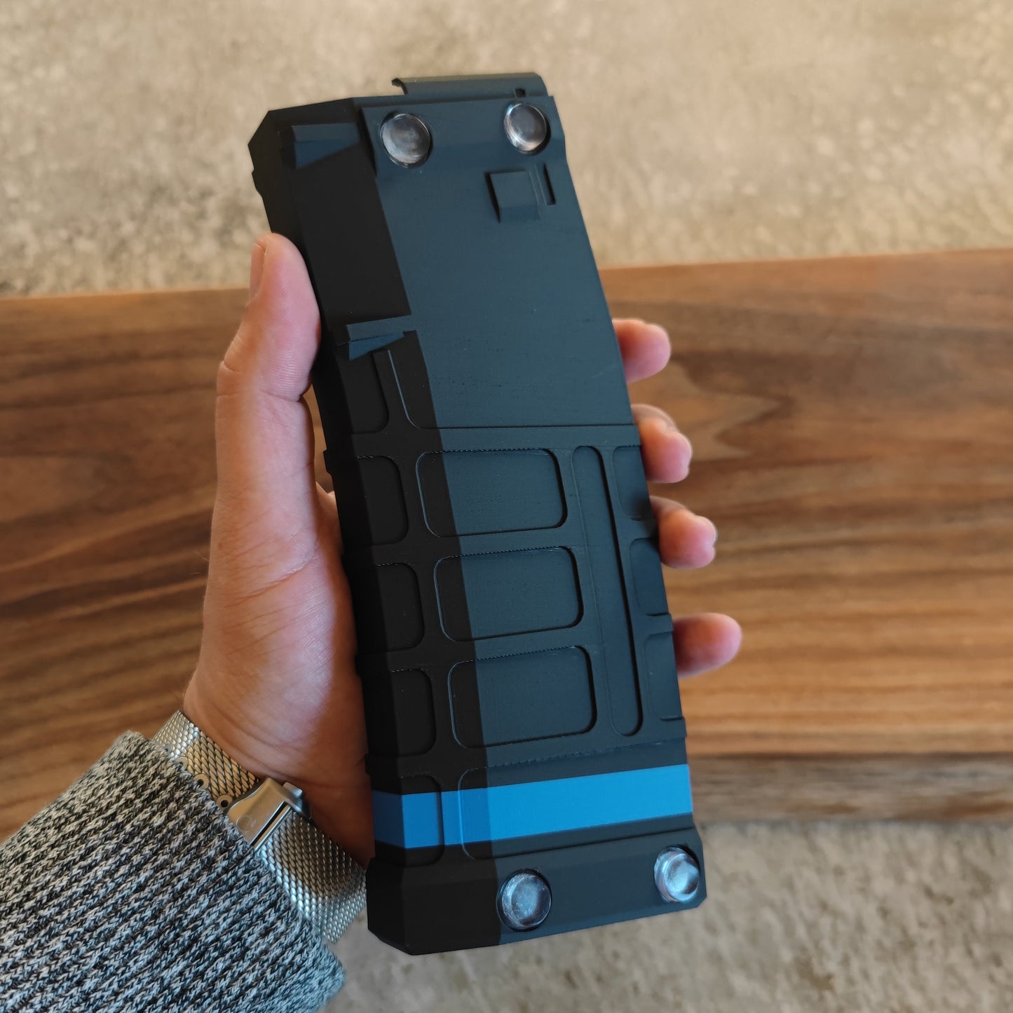 A hand holds the GEN4 CoinMag - Blue Line by coin-mag.net, a black and blue rectangular challenge coin holder with a rugged, textured surface and four metal circular accents, shown against a wooden background.