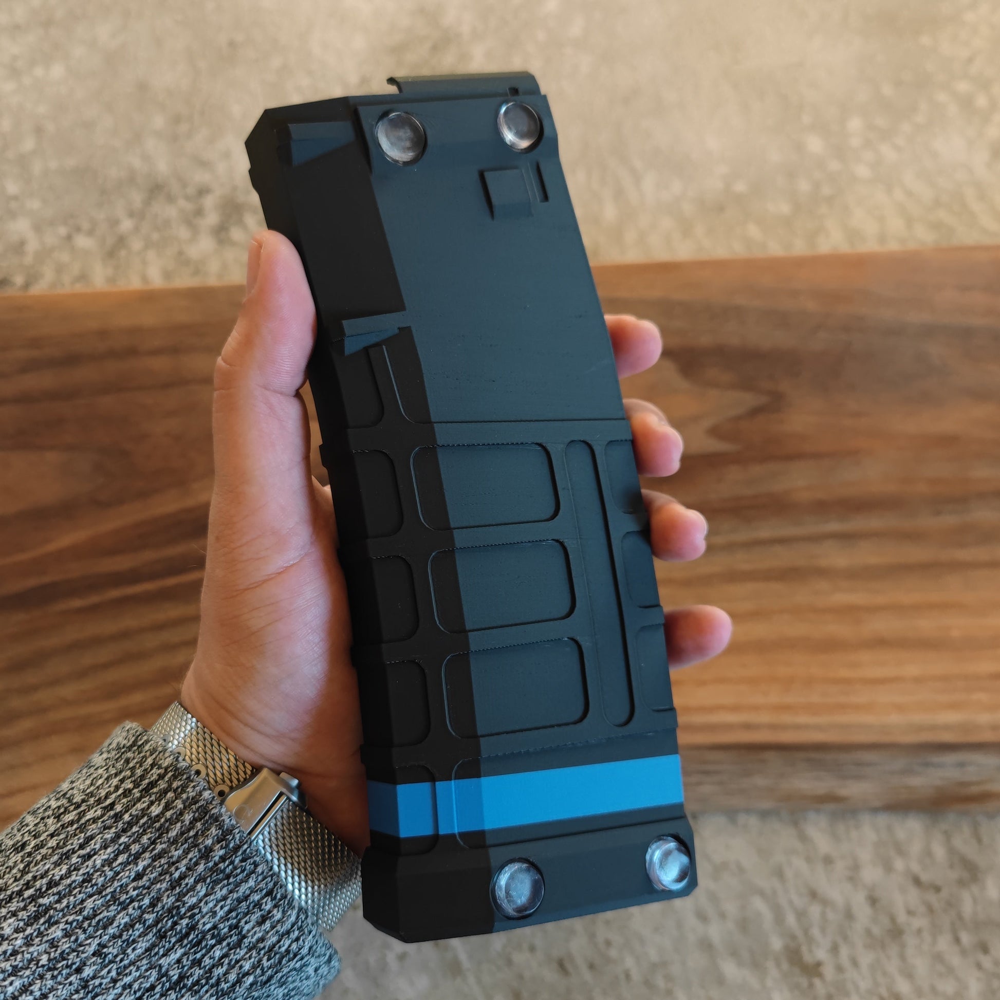 A hand holds the GEN4 CoinMag - Blue Line by coin-mag.net, a black and blue rectangular challenge coin holder with a rugged, textured surface and four metal circular accents, shown against a wooden background.
