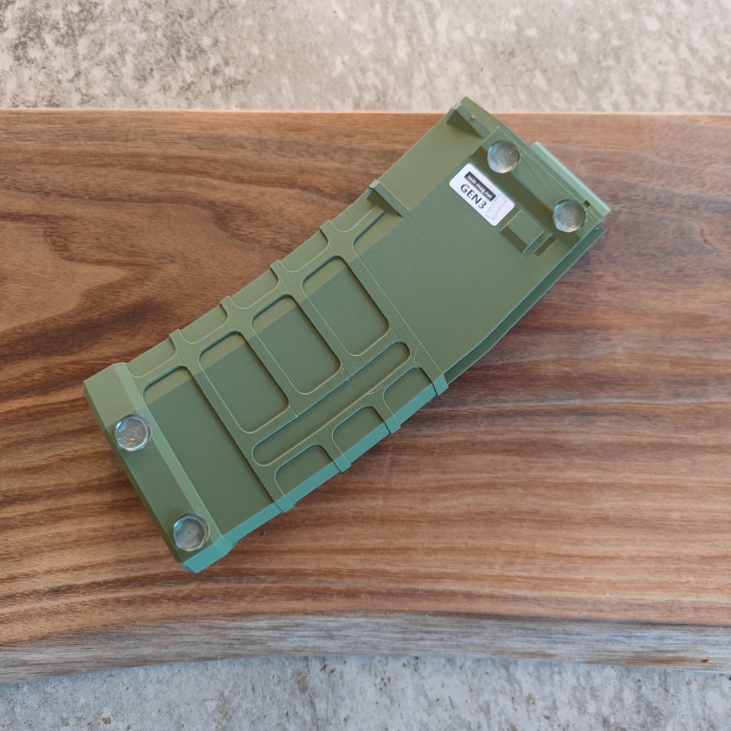 The GEN3 CoinMag - Olive by coin-mag.net, a green rectangular coin holder featuring an "OPEN" sticker and four screws, sits on a brown wooden surface with a lighter, textured background above.