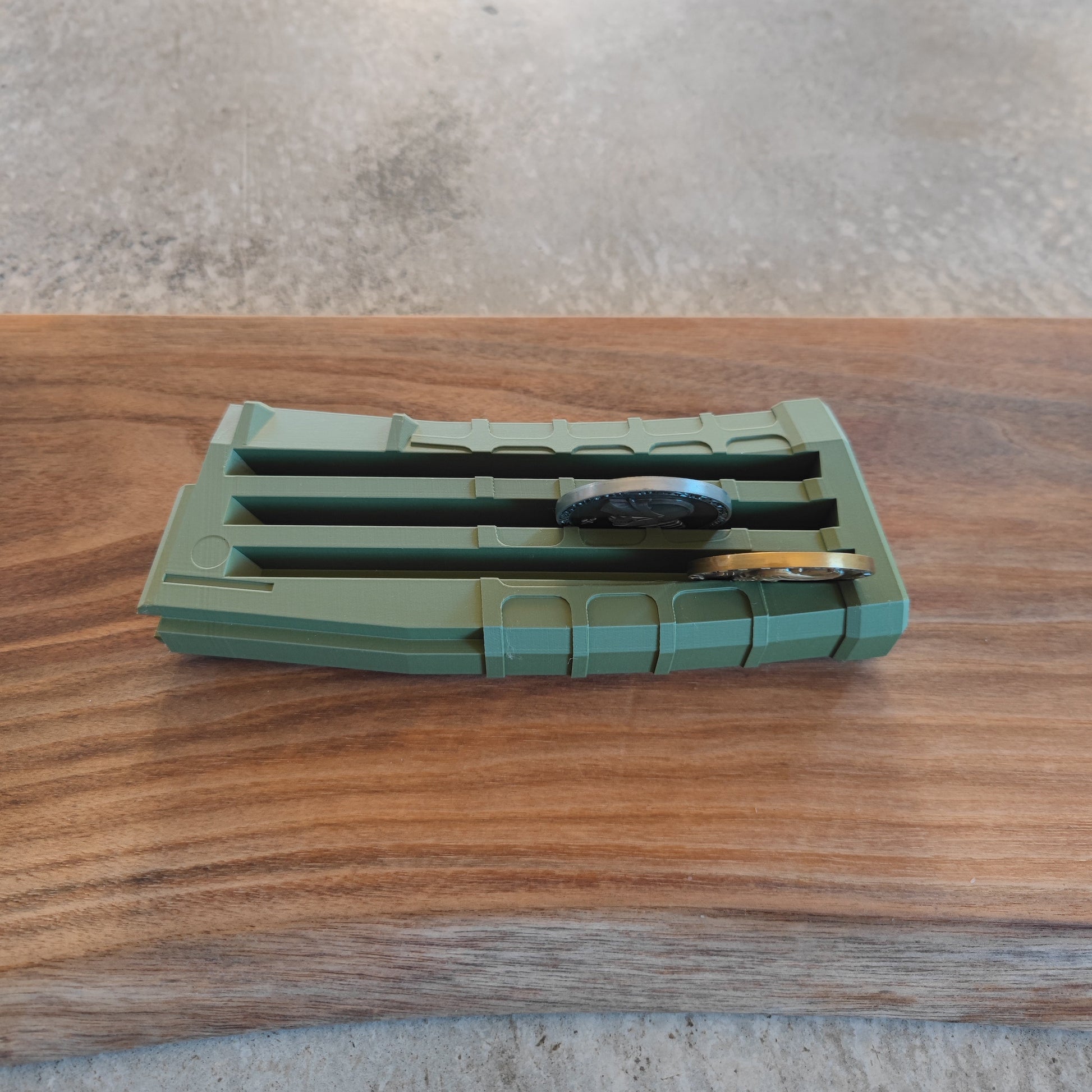The GEN3 CoinMag - Olive by coin-mag.net, a green rectangular coin holder with a slotted top, rests on wood and displays two rings—one black, one gold—against a gray textured backdrop.