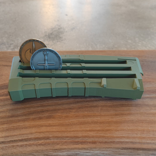 The GEN3 CoinMag - Olive by coin-mag.net is a green, rectangular holder with three slots. It displays two round metal tokens—a sword and a spear design—magazine-style on wood, set against a concrete background.