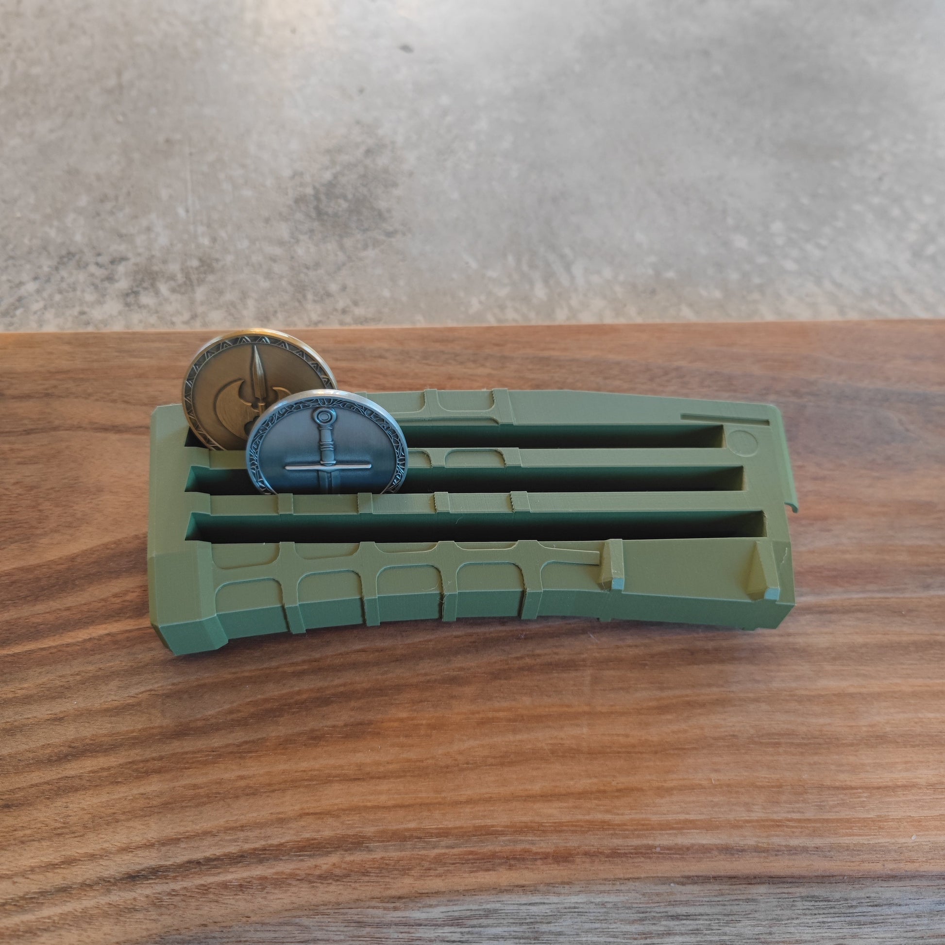 The GEN3 CoinMag - Olive by coin-mag.net, a green magazine-style coin display, sits on a wooden surface holding two metal coins upright in its slots against a gray, slightly textured background.