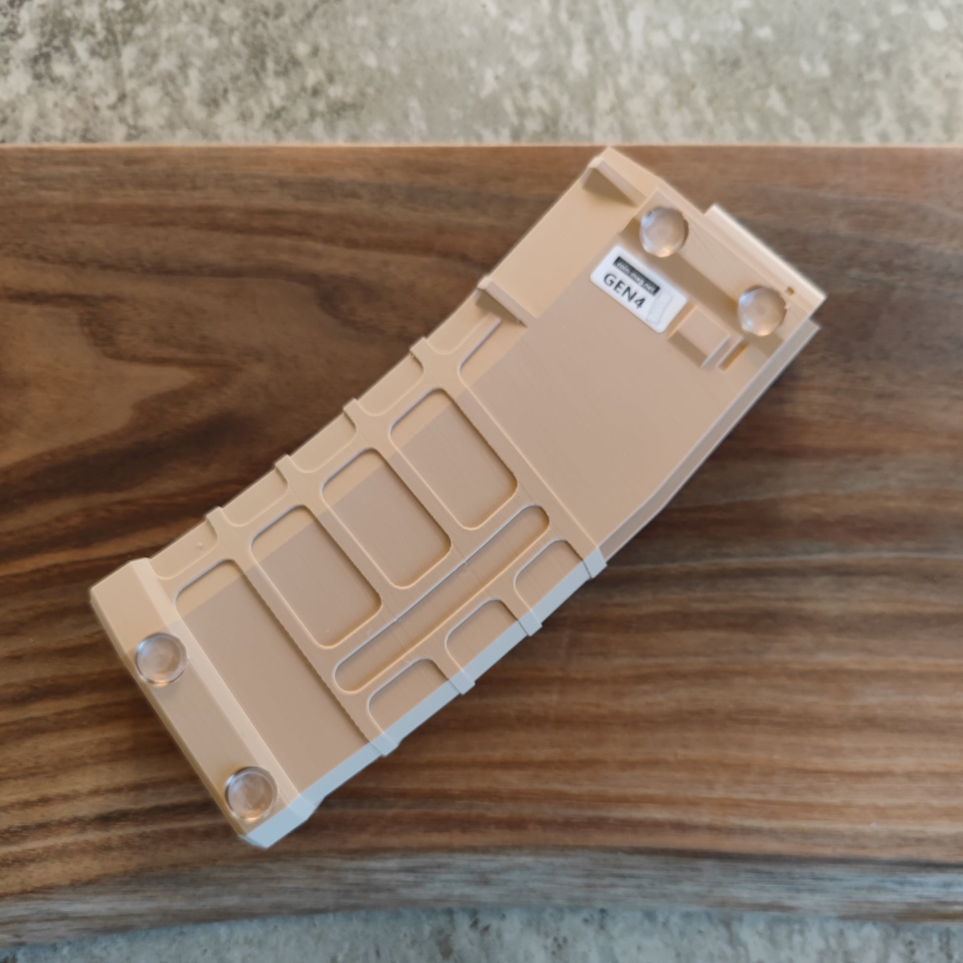 The GEN4 CoinMag - Sand by coin-mag.net is a tan, rectangular challenge coin holder with angled edges and clear feet, placed on wood. It features a GEN4 label and resembles a stylized, curved magazine or storage device.