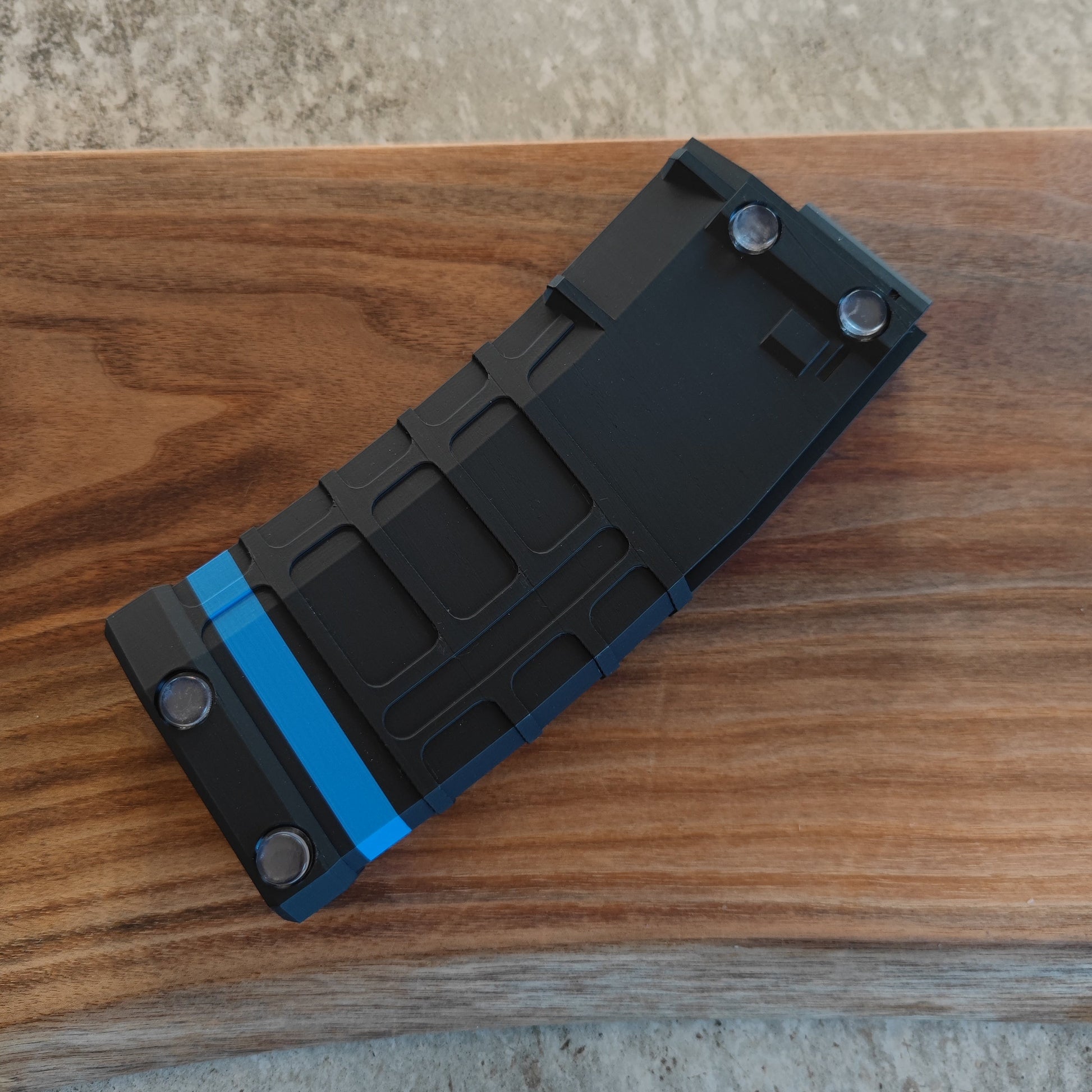 The GEN4 CoinMag - Blue Line by coin-mag.net, a black metal money clip with a blue stripe and silver screws, sits on polished wood next to a sleek challenge coin display.