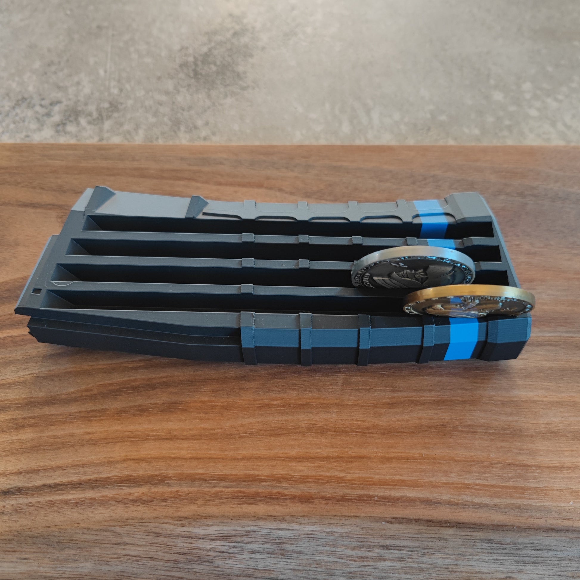 The GEN4 CoinMag - Blue Line by coin-mag.net is a black, ridged plastic holder with blue stripes that displays four military coins upright on a wooden surface, blurred background included.