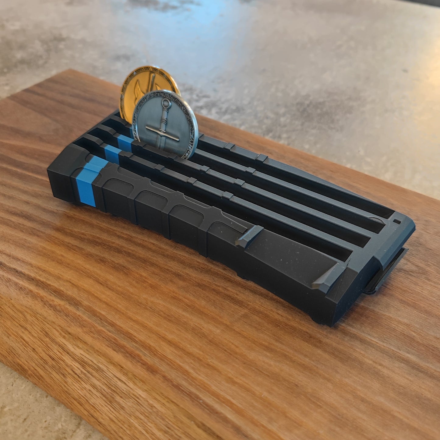 The GEN4 CoinMag - Blue Line by coin-mag.net is a black, rectangular holder with blue accents that displays two coins upright on a wooden base, shown against a concrete background.