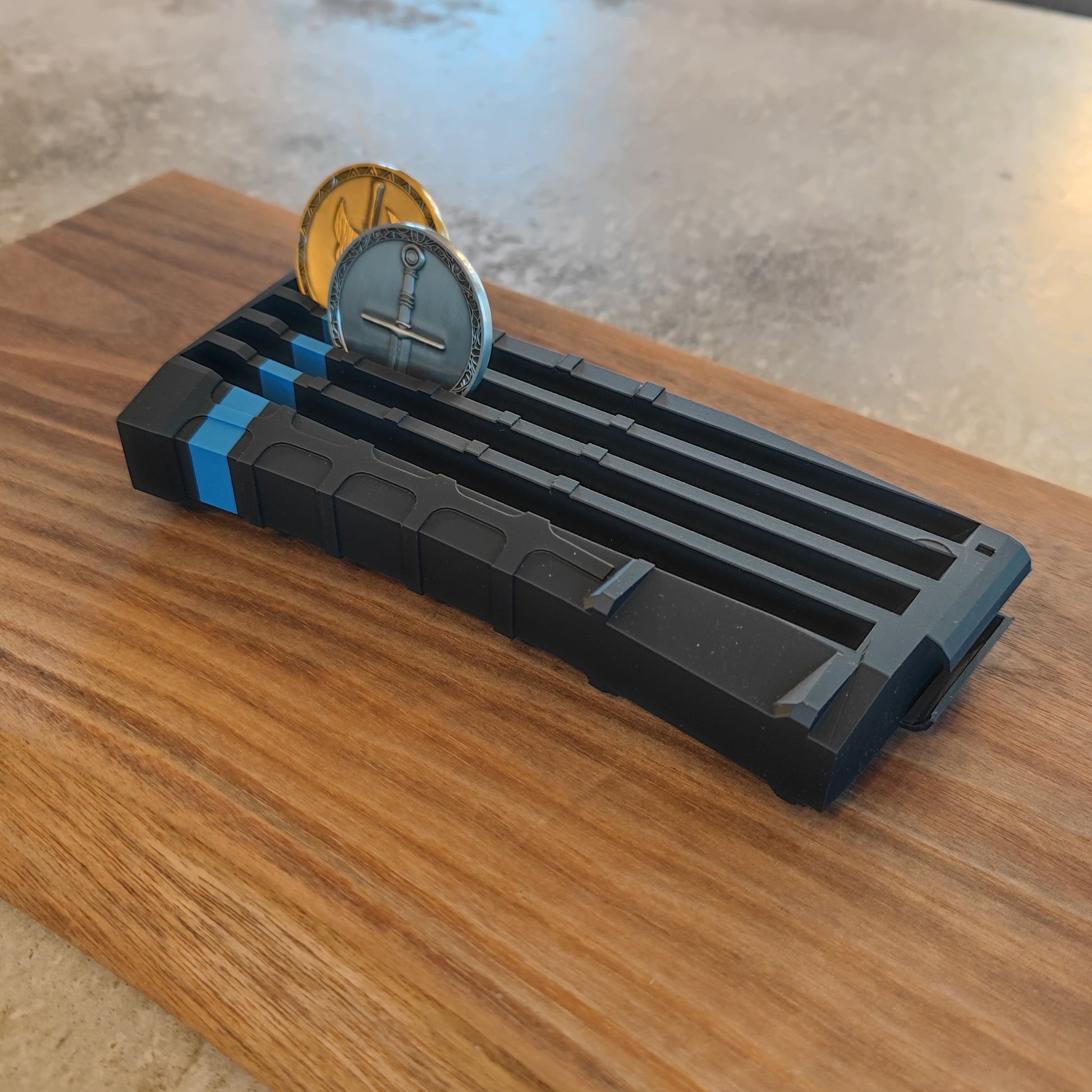 The GEN4 CoinMag - Blue Line by coin-mag.net is a black, rectangular holder with blue accents that displays two coins upright on a wooden base, shown against a concrete background.