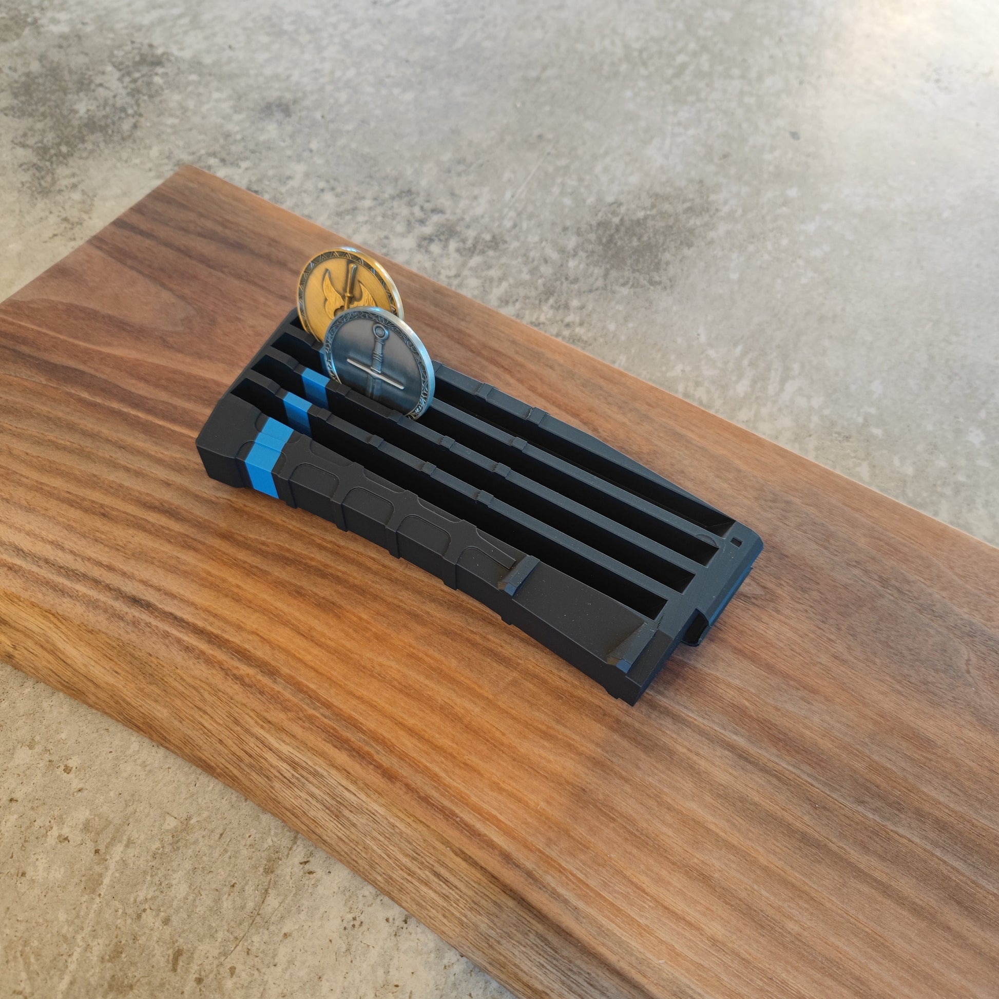 The GEN4 CoinMag - Blue Line by coin-mag.net, a black and blue challenge coin holder with two coins inserted, rests on a rectangular wooden board placed on concrete.