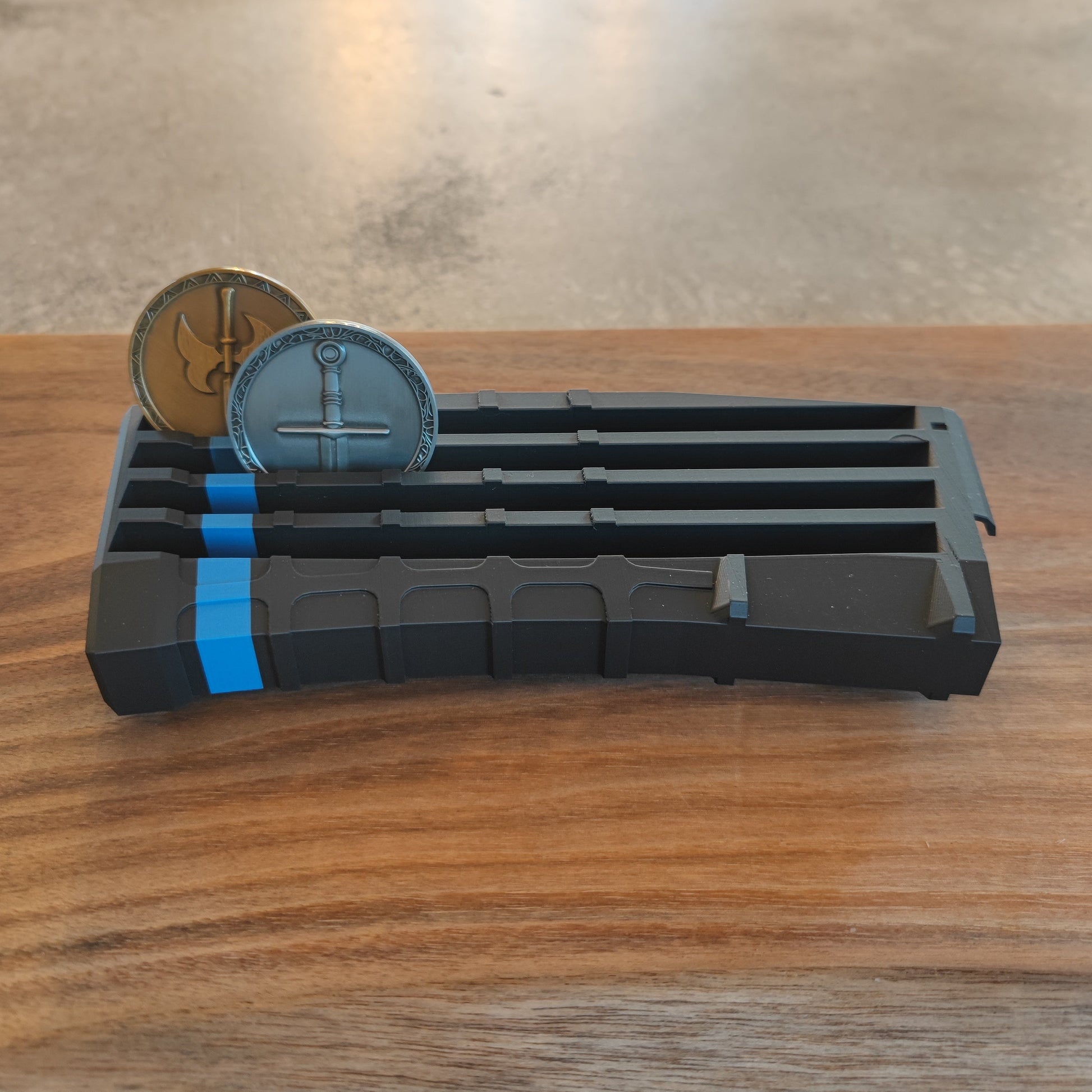 The GEN4 CoinMag - Blue Line by coin-mag.net is a black, rectangular holder with blue accents that displays two round coins upright in separate slots. It’s shown on a wooden surface with a blurred gray background.