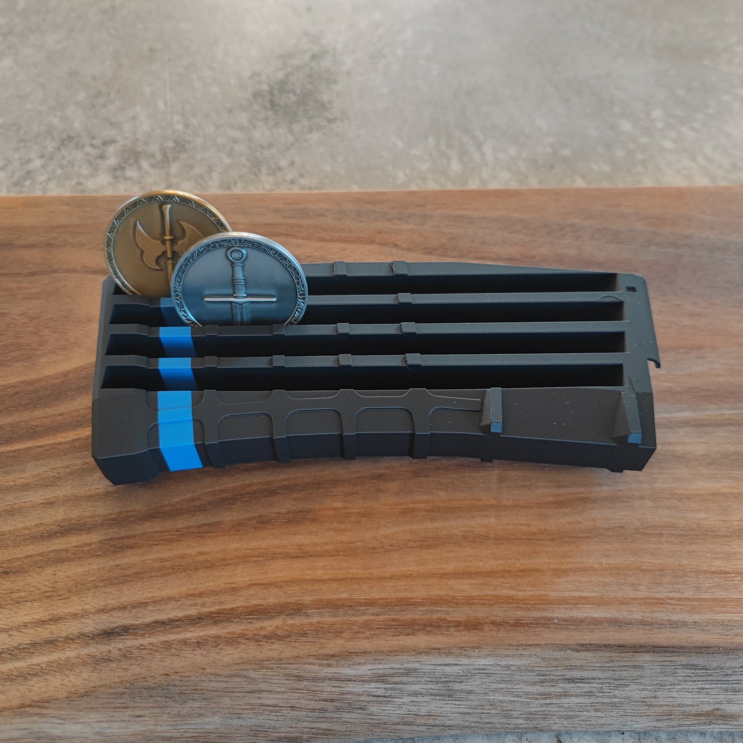 The GEN4 CoinMag - Blue Line by coin-mag.net, a black and blue plastic challenge coin holder, rests on a wooden surface with two metallic coins featuring anchor designs partially inserted in separate slots.