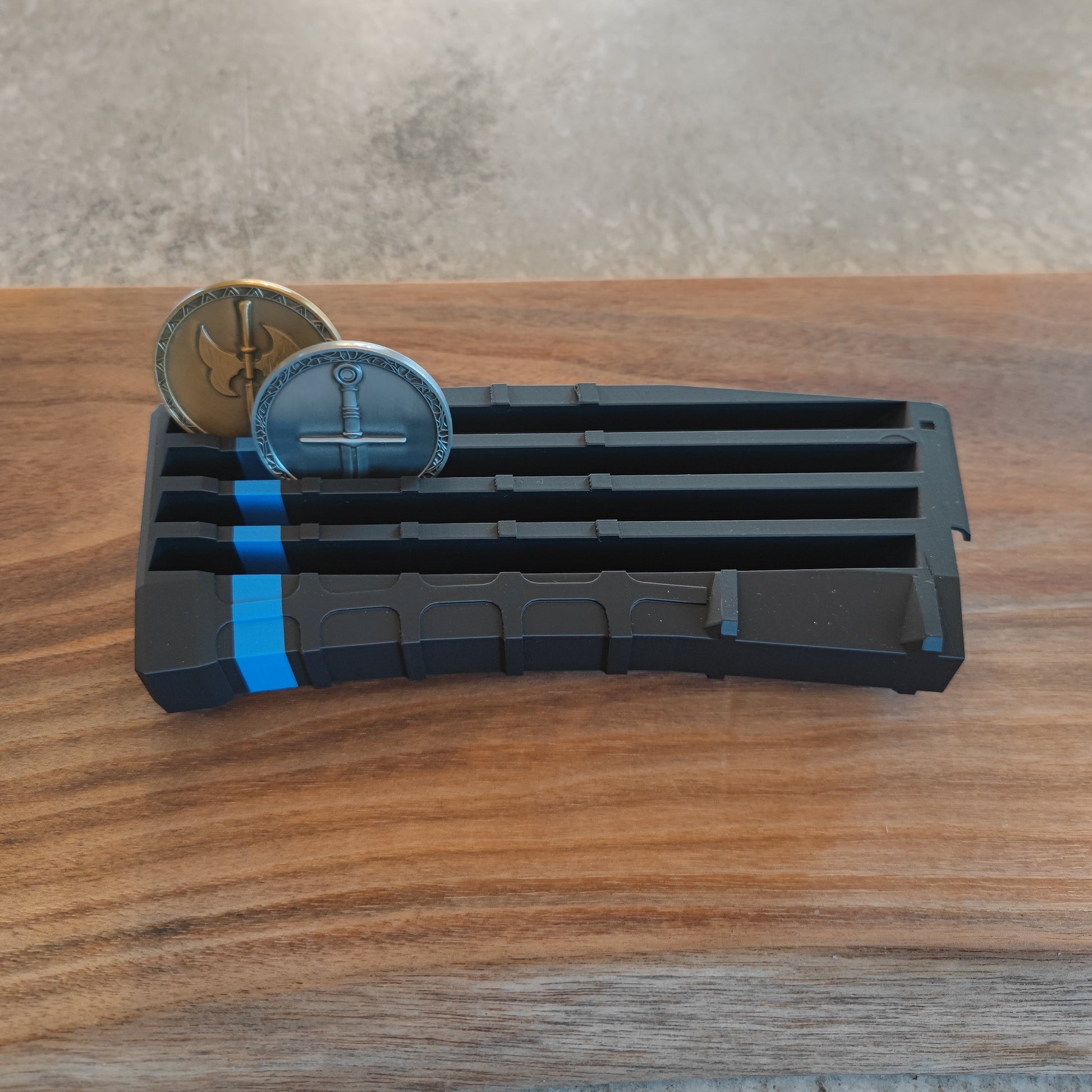 The GEN4 CoinMag - Blue Line by coin-mag.net, a black and blue plastic challenge coin holder, rests on a wooden surface with two metallic coins featuring anchor designs partially inserted in separate slots.