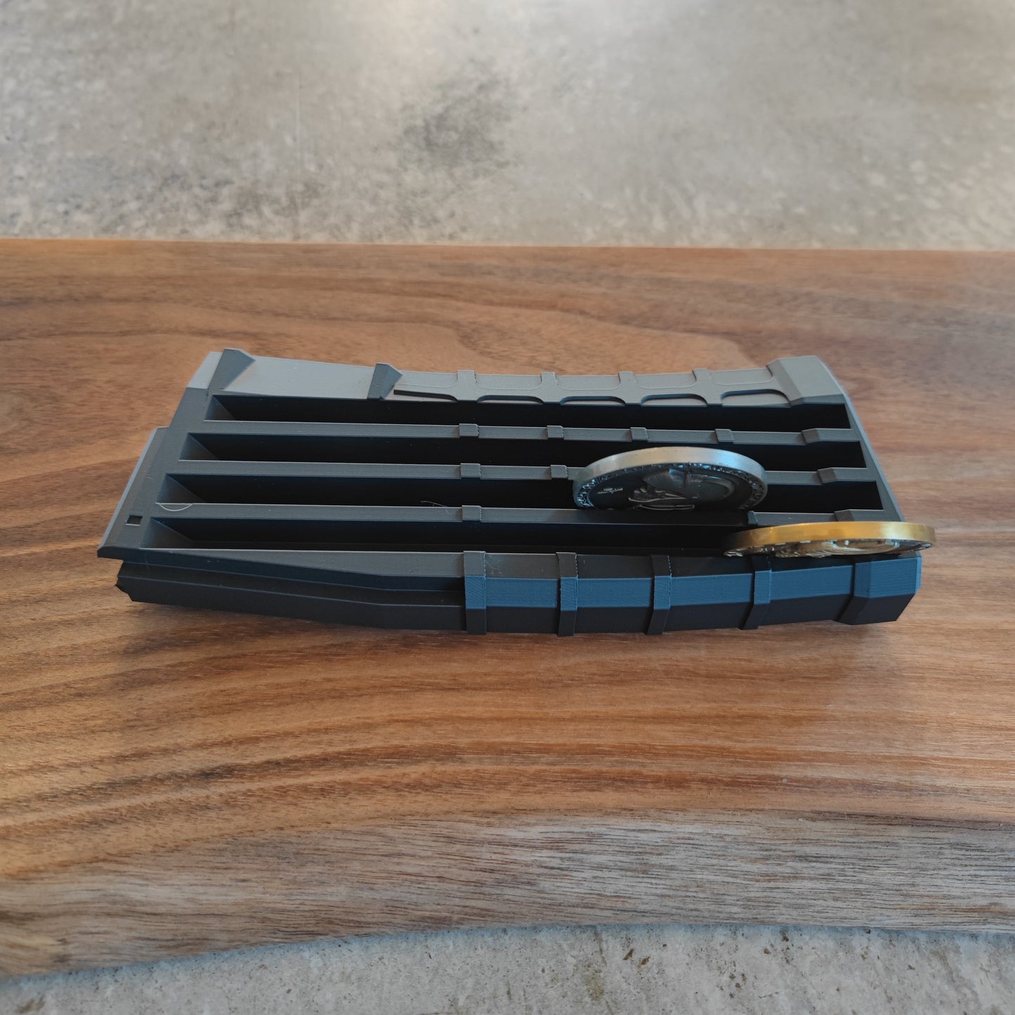 The GEN4 CoinMag - Black by coin-mag.net is a slotted, black holder that displays two watches or challenge coins securely on a wood surface with angled grooves to keep items neatly in place.