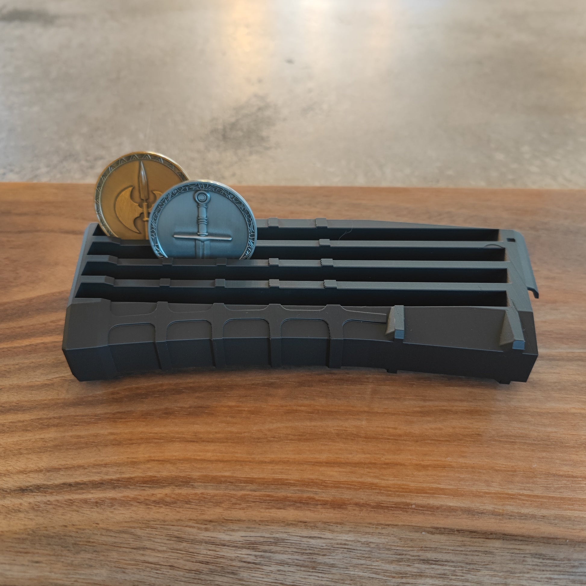 The GEN4 CoinMag - Black by coin-mag.net, a rectangular challenge coin holder with slots, displays two sword-themed metal coins on a wooden surface. The background appears blurred.