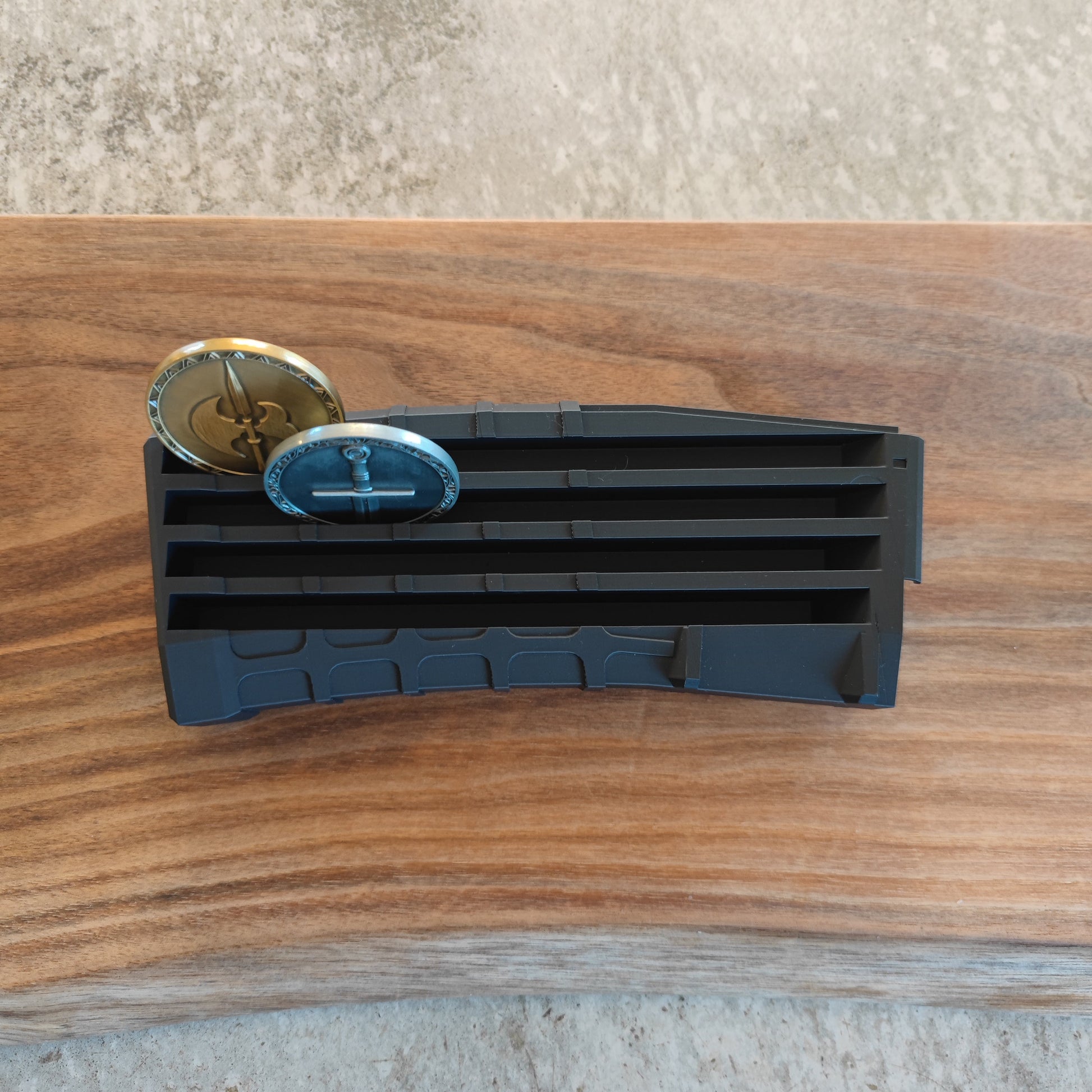 The GEN4 CoinMag - Black by coin-mag.net, a black plastic challenge coin holder shaped like a magazine, holds two metal challenge coins in its top slot. It sits on wood, with a partially visible concrete background.