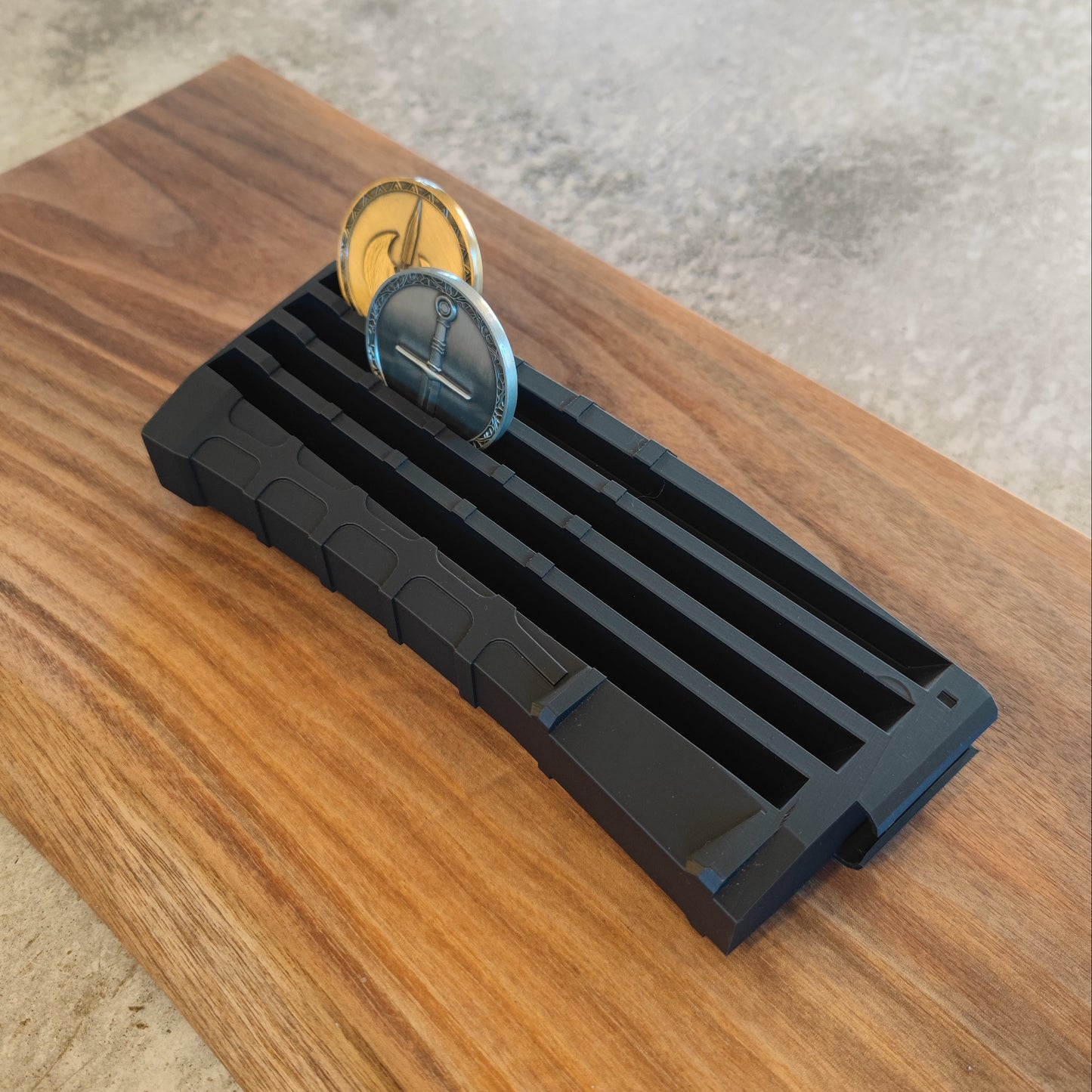 The GEN4 CoinMag - Black by coin-mag.net, a ridged magazine-shaped challenge coin holder, displays two upright coins in its grooves on a wooden surface against a concrete-textured background.
