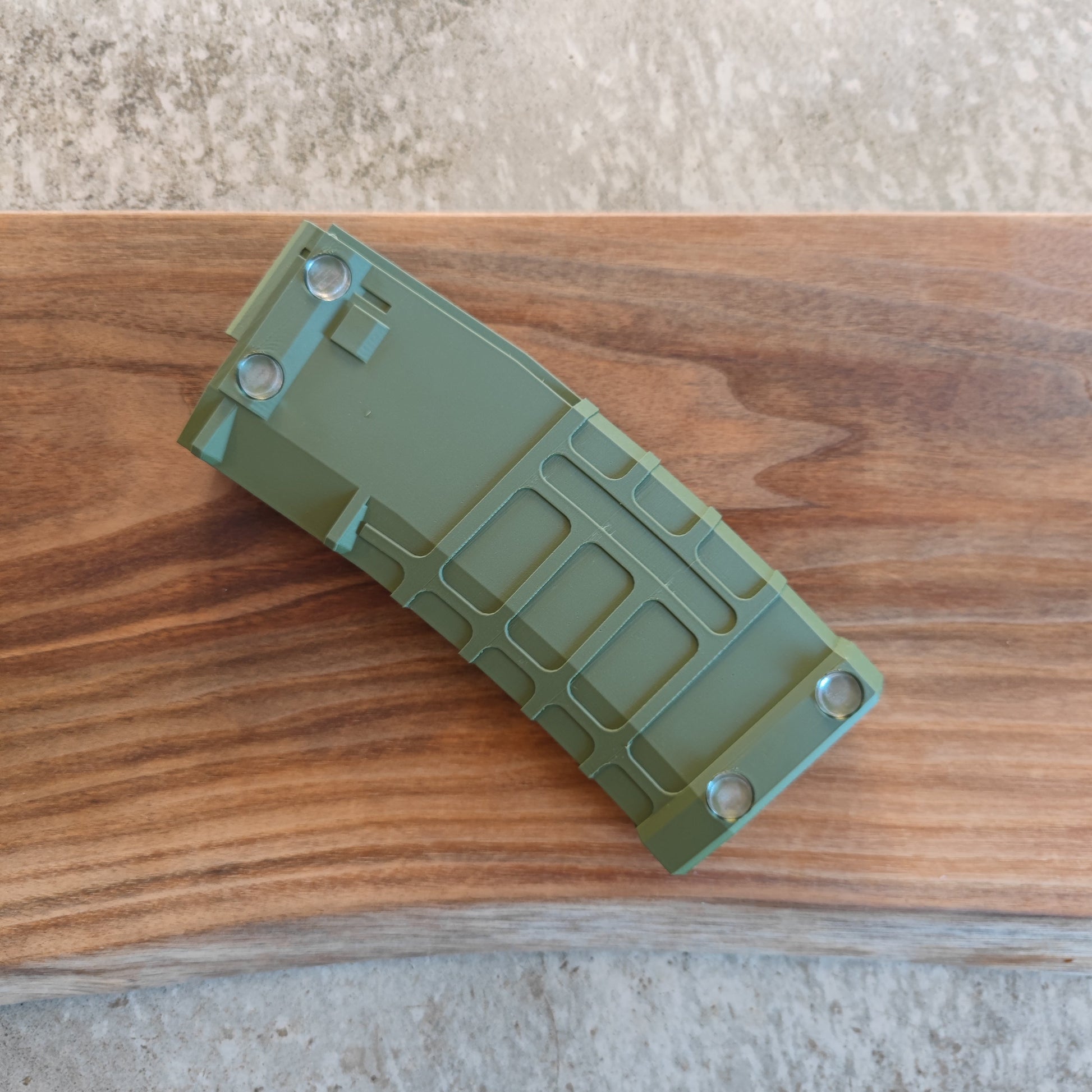 The GEN4 CoinMag - Olive by coin-mag.net, a green rectangular coin holder with grooves and four metal corner accents, rests on a wooden surface with natural grain. A concrete floor appears in the background.