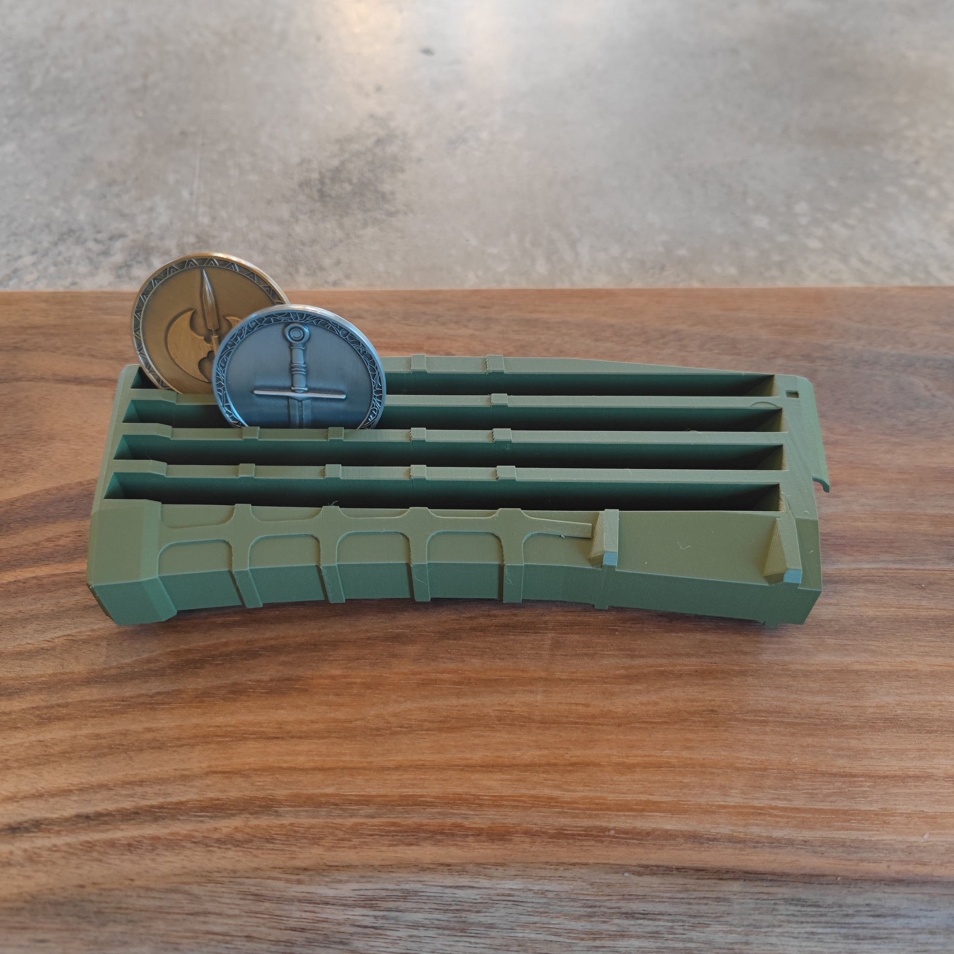 The GEN4 CoinMag - Olive by coin-mag.net is a green, rectangular coin holder with four grooves that displays two round coins upright on a wooden surface, making for an attractive challenge coin presentation.