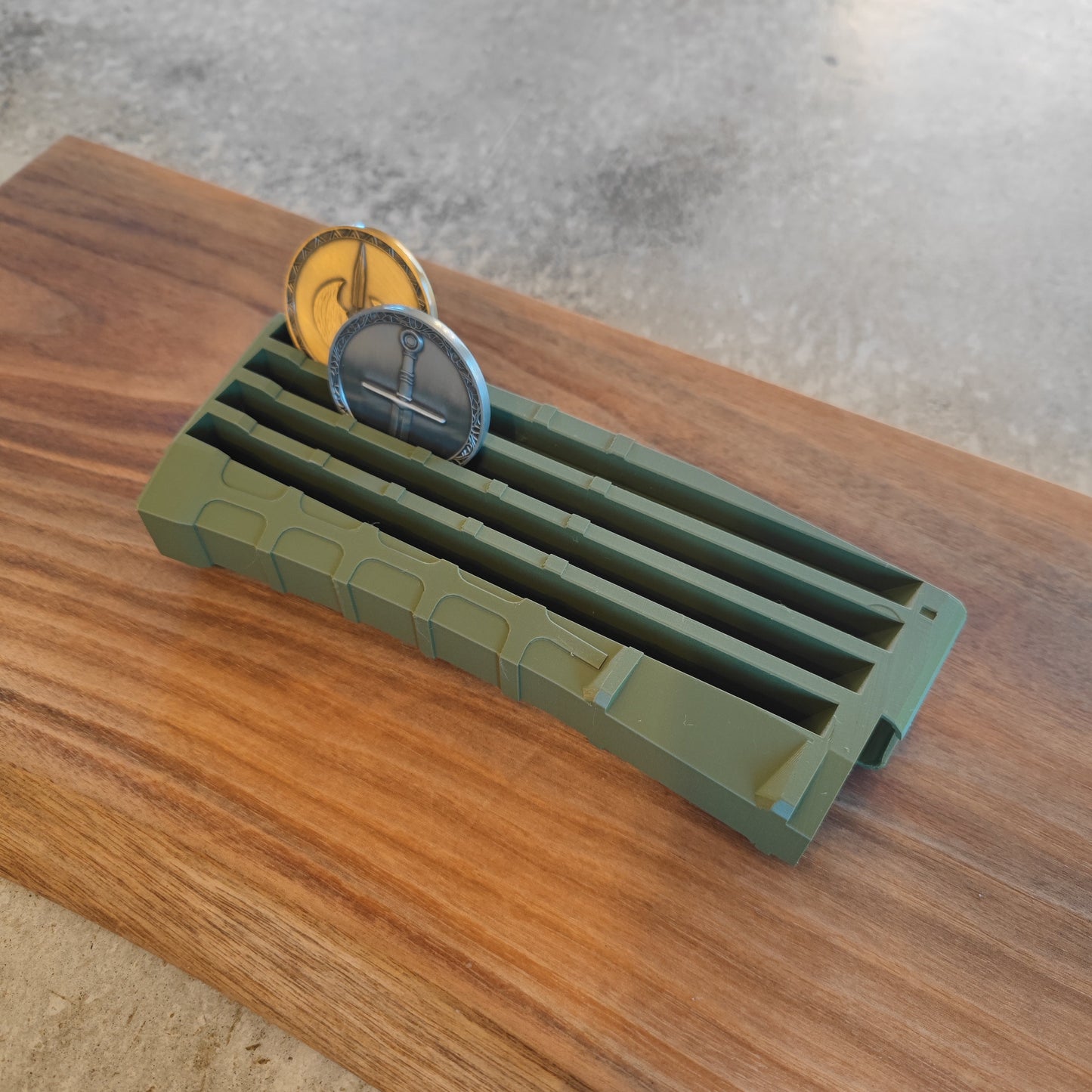 The coin-mag.net GEN4 CoinMag - Olive, a rifle magazine-shaped coin display, holds two challenge coins on a wooden surface with a concrete floor in the background.