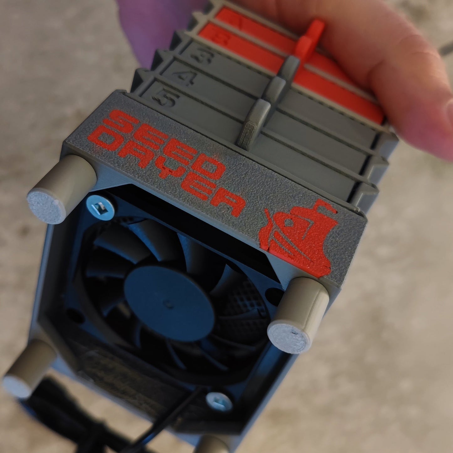 A hand holds the Seed Dryer - Classic Console by PepperMerchant.net, a gray and red USB-powered seed dryer with a labeled fan, numbered stacked trays, and a 3D printer-style icon—showcasing modern seed saving technology.