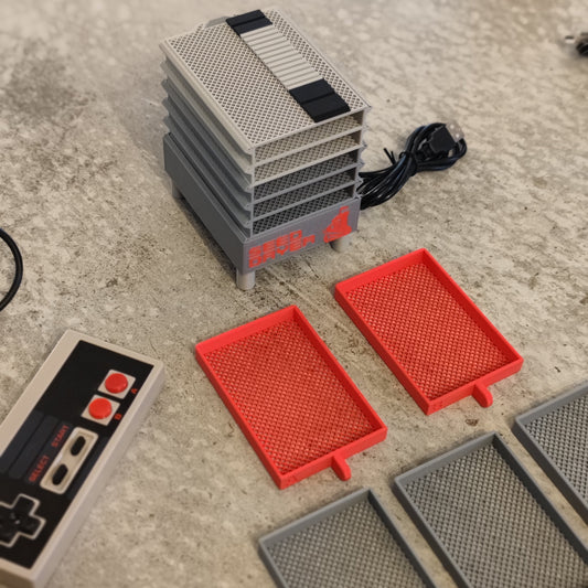A stack of gray and red grid-patterned trays, a black cable, and a classic Nintendo-style controller on a textured surface—PepperMerchant.net’s Seed Dryer - Classic Console for innovative seed drying.