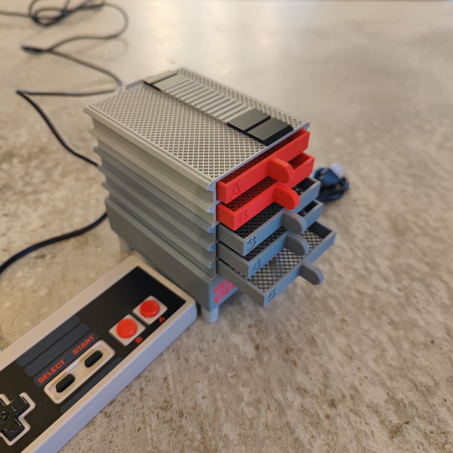 A close-up of PepperMerchant.net’s Seed Dryer - Classic Console, styled like NES consoles with small gray and red drawers—ideal for seed saving technology—next to a classic NES controller on a textured surface.