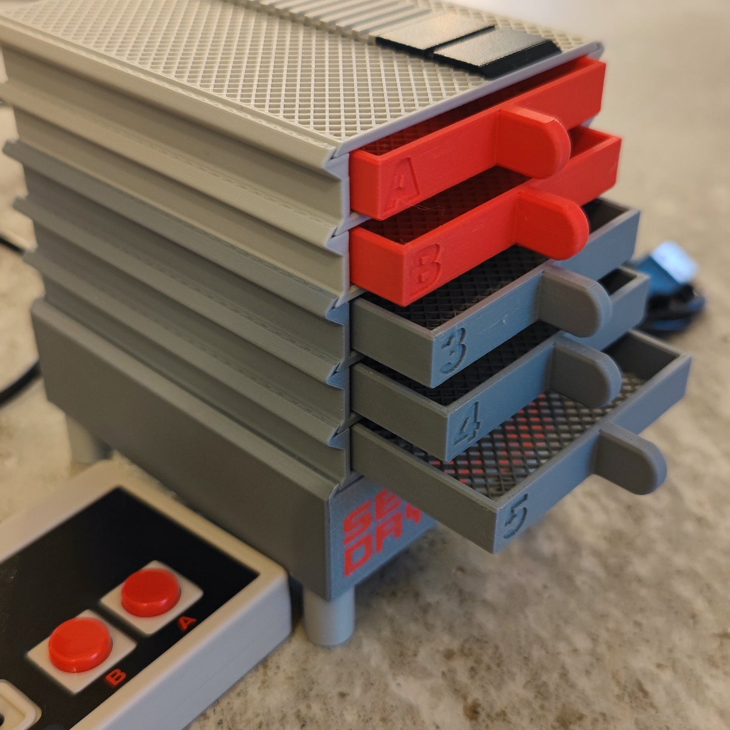 A stackable storage rack with red "A" and "B" tabs and gray trays numbered "3," "4," and "5" is shown. Nearby is the Seed Dryer - Classic Console by PepperMerchant.net, next to a retro-style game controller, highlighting modern seed saving.
