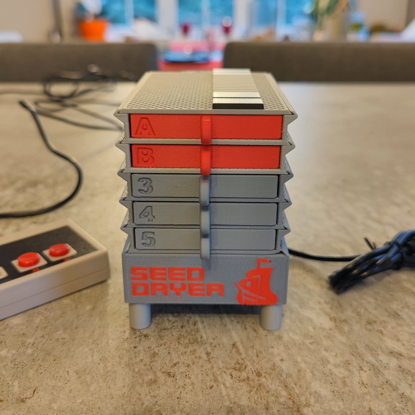 The PepperMerchant.net Seed Dryer - Classic Console is a compact, gray unit with five stacked trays—top two marked "A" and "B" in red, others numbered "3," "4," and "5." An electronic controller sits to the left on a gray tabletop.
