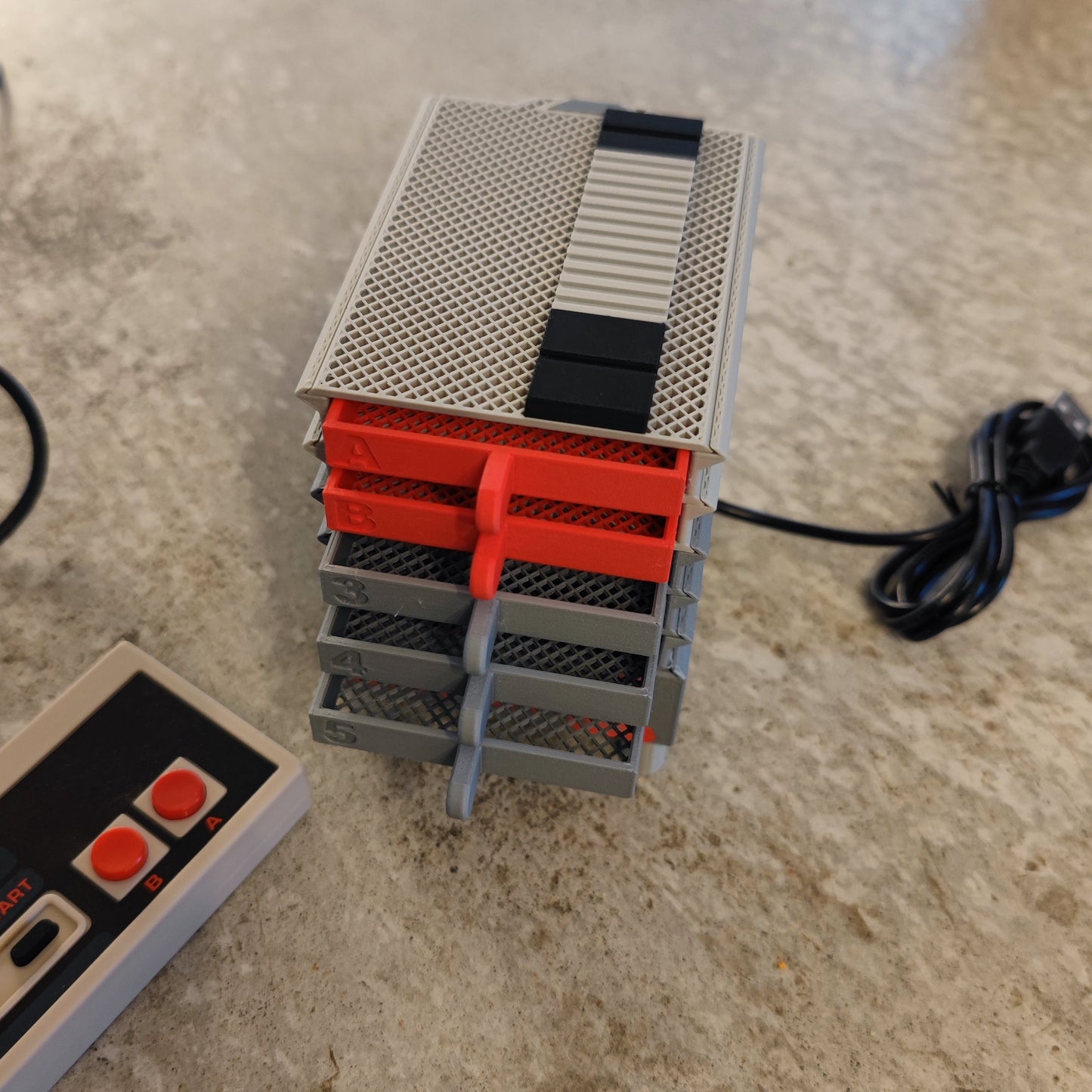 The Seed Dryer - Classic Console from PepperMerchant.net, featuring gray and red trays with a USB cable, sits beside a retro game controller—highlighting modern seed-saving innovation for today’s gardeners.