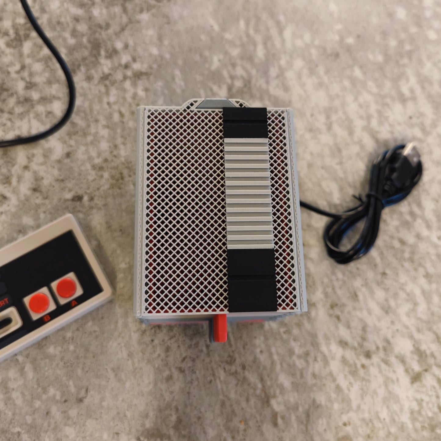 A retro video game controller with red buttons lies next to the PepperMerchant.net Seed Dryer - Classic Console—an innovative, USB-powered seed saver with black and silver stripes, a coiled cable, and a textured surface.