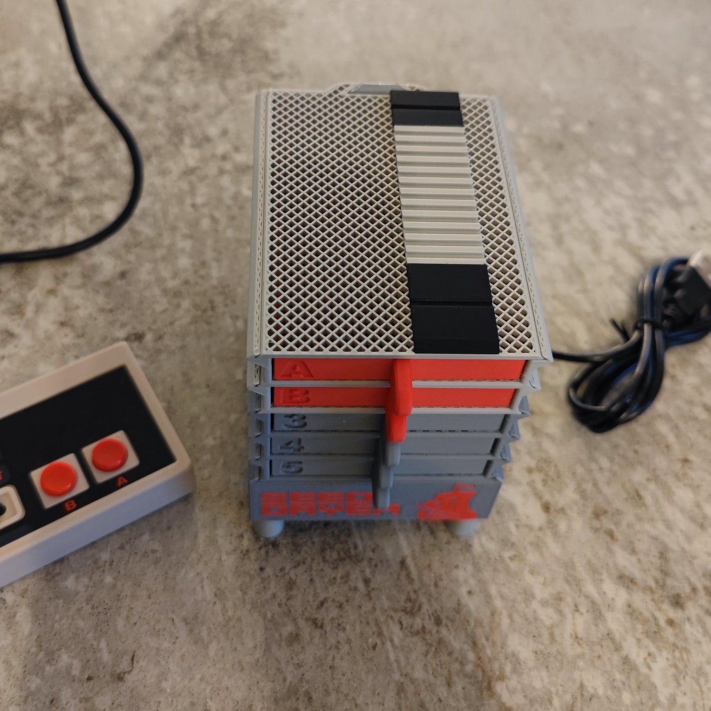 A close-up of the PepperMerchant.net Seed Dryer - Classic Console, a retro-style gray and red device resembling stacked boxes, with a USB-powered seed dryer and rectangular controller resting on a textured surface.