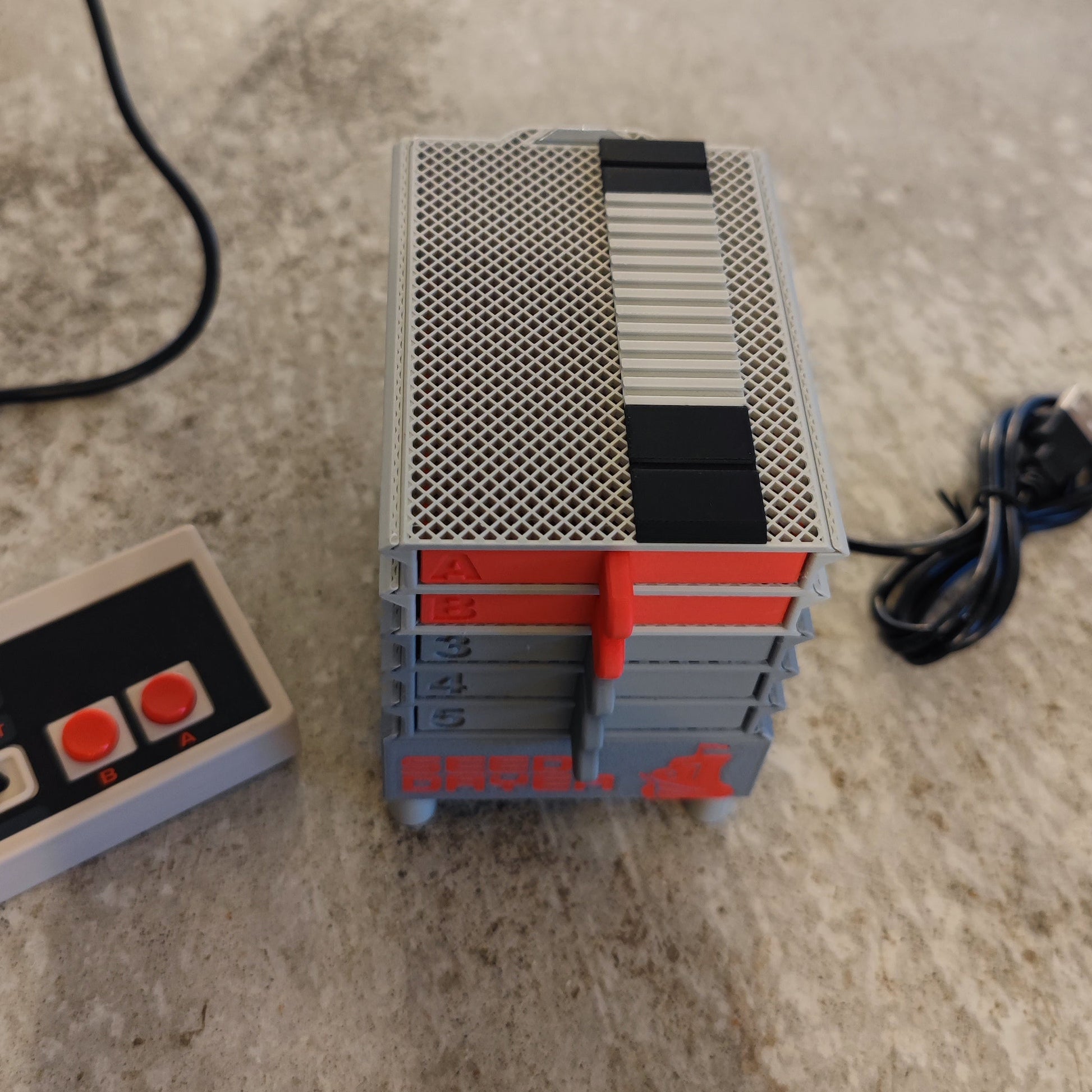 A close-up of the PepperMerchant.net Seed Dryer - Classic Console, a retro-style gray and red device resembling stacked boxes, with a USB-powered seed dryer and rectangular controller resting on a textured surface.