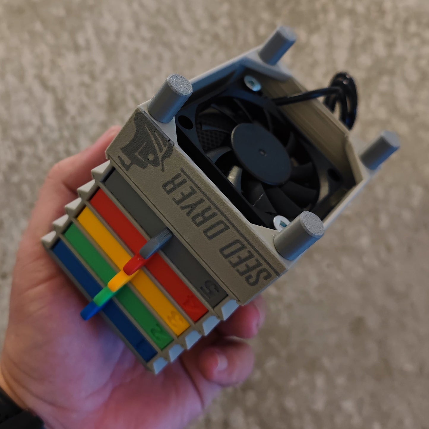 A hand holds the Seed Dryer - Super Classic Console by PepperMerchant.net, a USB-powered device with a top fan and vibrant stacked compartments in yellow, green, blue, orange, and red—showcasing modern seed saving technology.