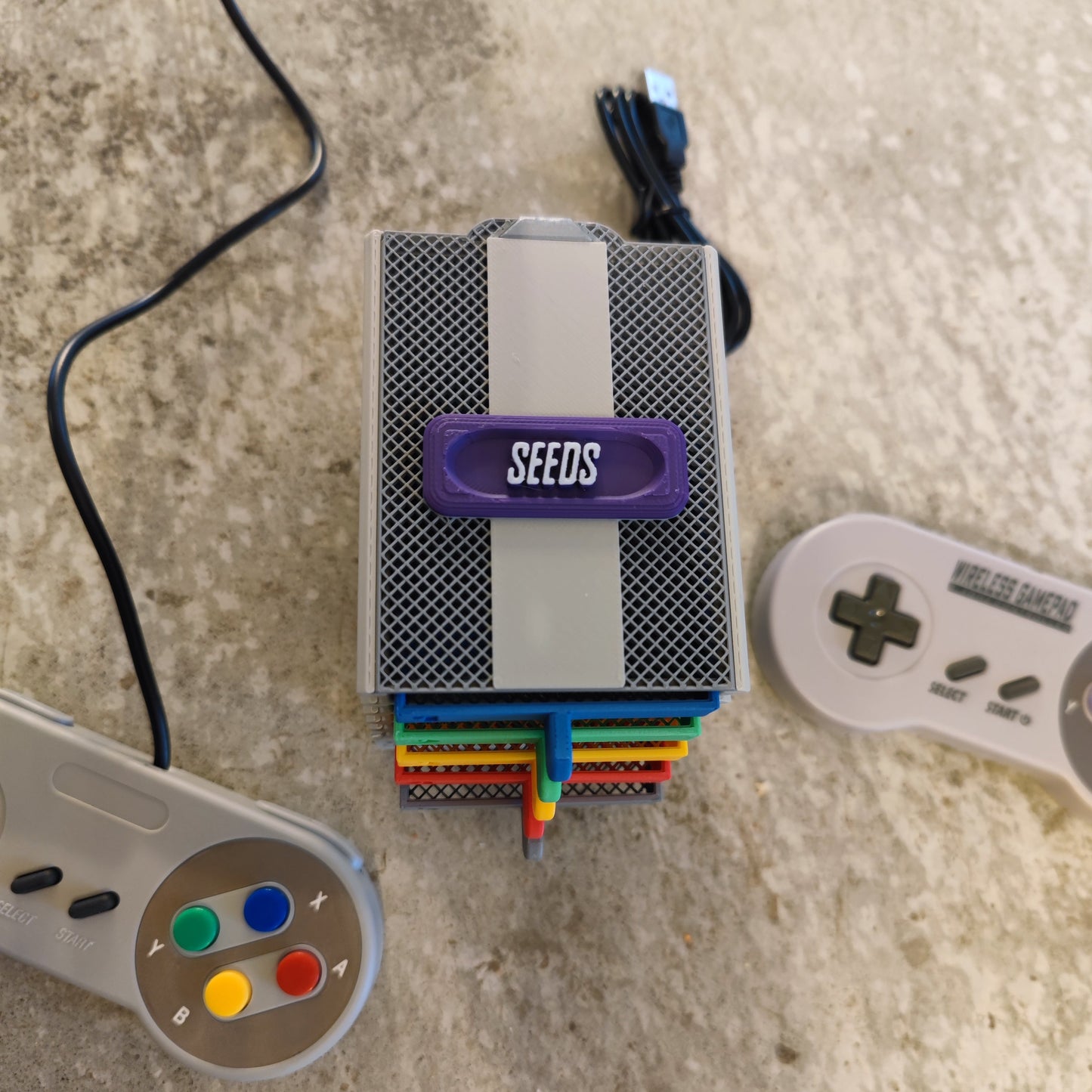 A compact, box-shaped Seed Dryer - Super Classic Console by PepperMerchant.net with heatless seed dryer technology and colorful cables sits on a textured surface, flanked by two retro game controllers.