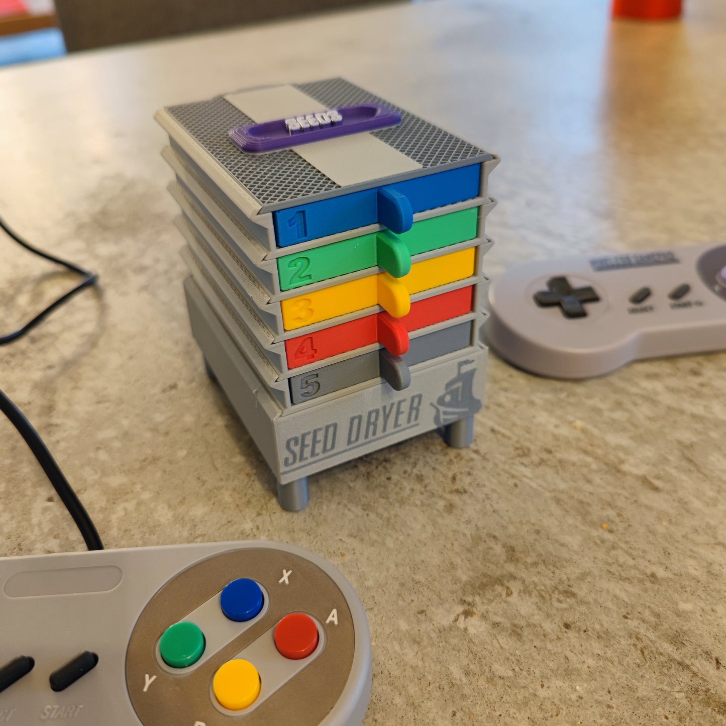 The Seed Dryer - Super Classic Console by PepperMerchant.net is a compact, gray, stackable USB-powered seed dryer with five numbered trays. Retro video game controllers add a playful, modern twist to your seed saving routine.