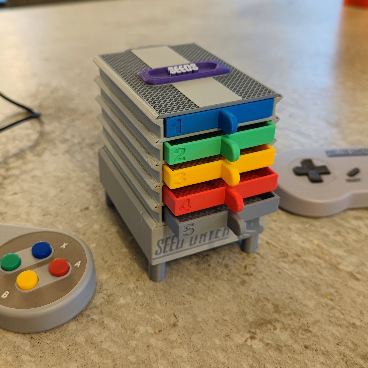 A stack of five cartridges labeled 1–5 in gray, blue, green, yellow, and red evokes a retro console. Nearby are two gray controllers with colorful buttons and PepperMerchant.net’s Seed Dryer – Super Classic Console for seed saving.