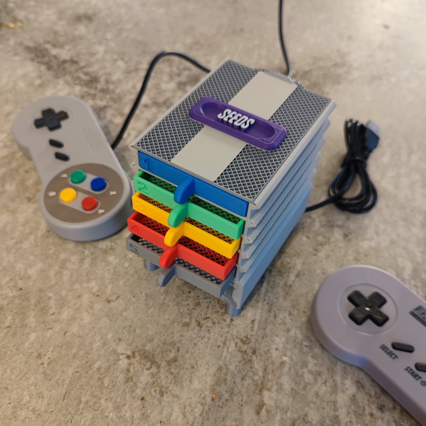 The Seed Dryer - Super Classic Console by PepperMerchant.net, styled like a mini SNES with stacked colored cartridges, sits on a table beside two gray retro controllers—one unwired and one coiled—displaying modern seed saving technology.