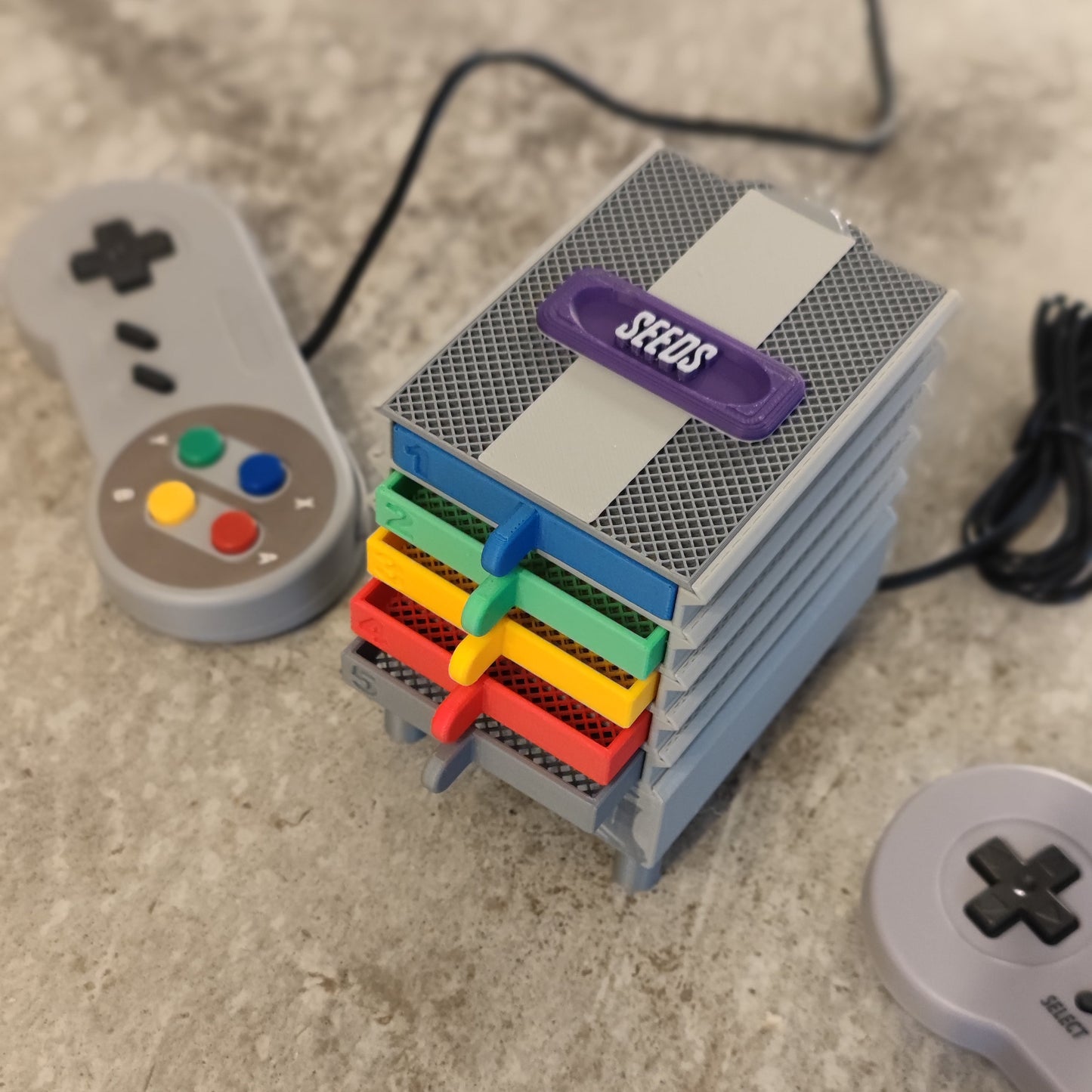 The PepperMerchant.net Seed Dryer - Super Classic Console sits on a table, USB-powered, with "SEEDS" labeled slots filled by colorful packets. Two classic game controllers—one colored, one gray—rest nearby.