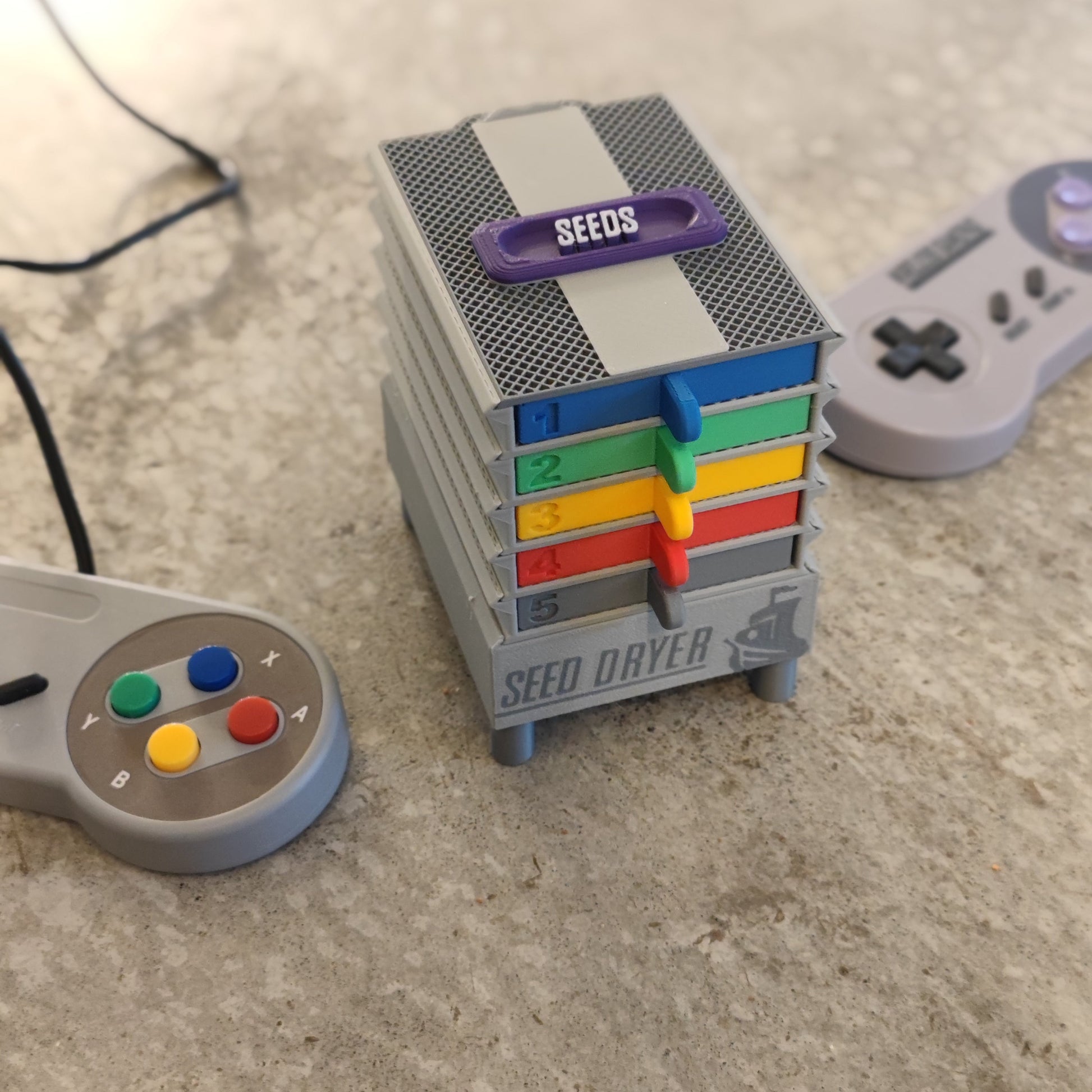 The PepperMerchant.net Seed Dryer - Super Classic Console, a compact gray USB-powered unit with five colorful numbered drawers, sits on the counter. Two retro game controllers and a "SEEDS" container rest nearby.