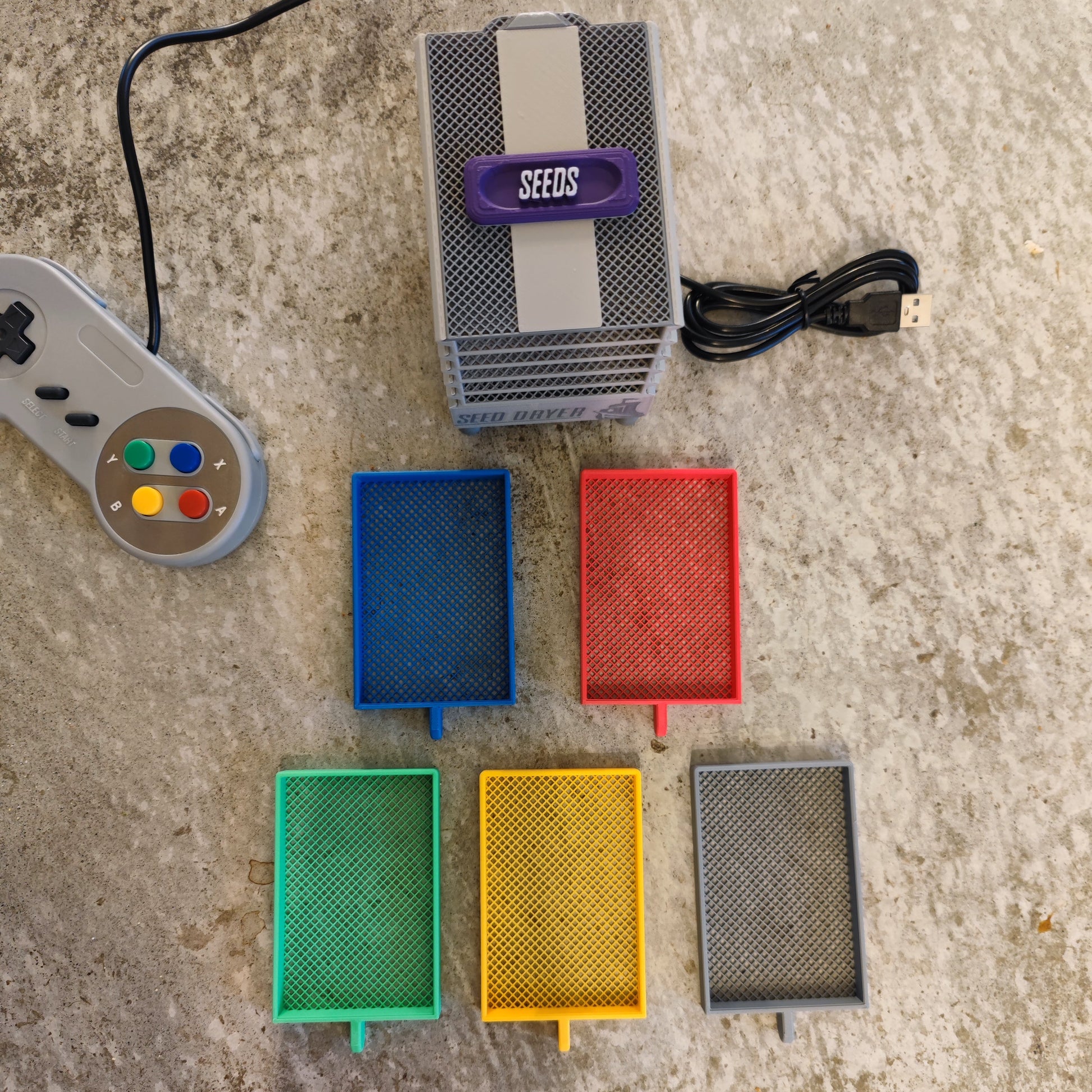 The PepperMerchant.net Seed Dryer - Super Classic Console is a gray device labeled "SEEDS" with a USB cable, game controller, and five colorful mesh trays (blue, red, green, yellow, gray) displayed on concrete—offering innovative seed saving technology.